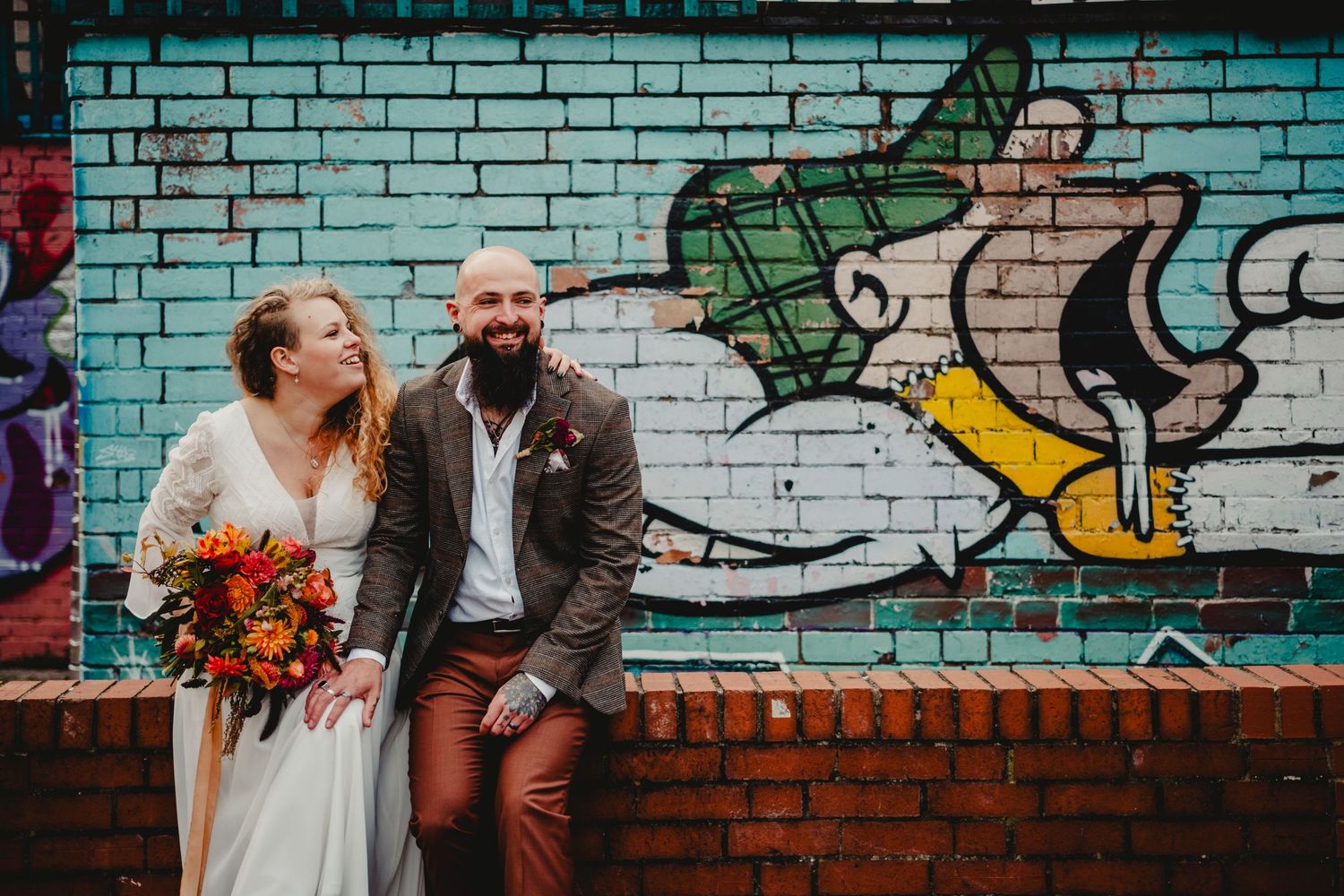 Newlywed couple smiles and sits against colorful brick wall with vibrant graffiti art and orange bridal flowers.
