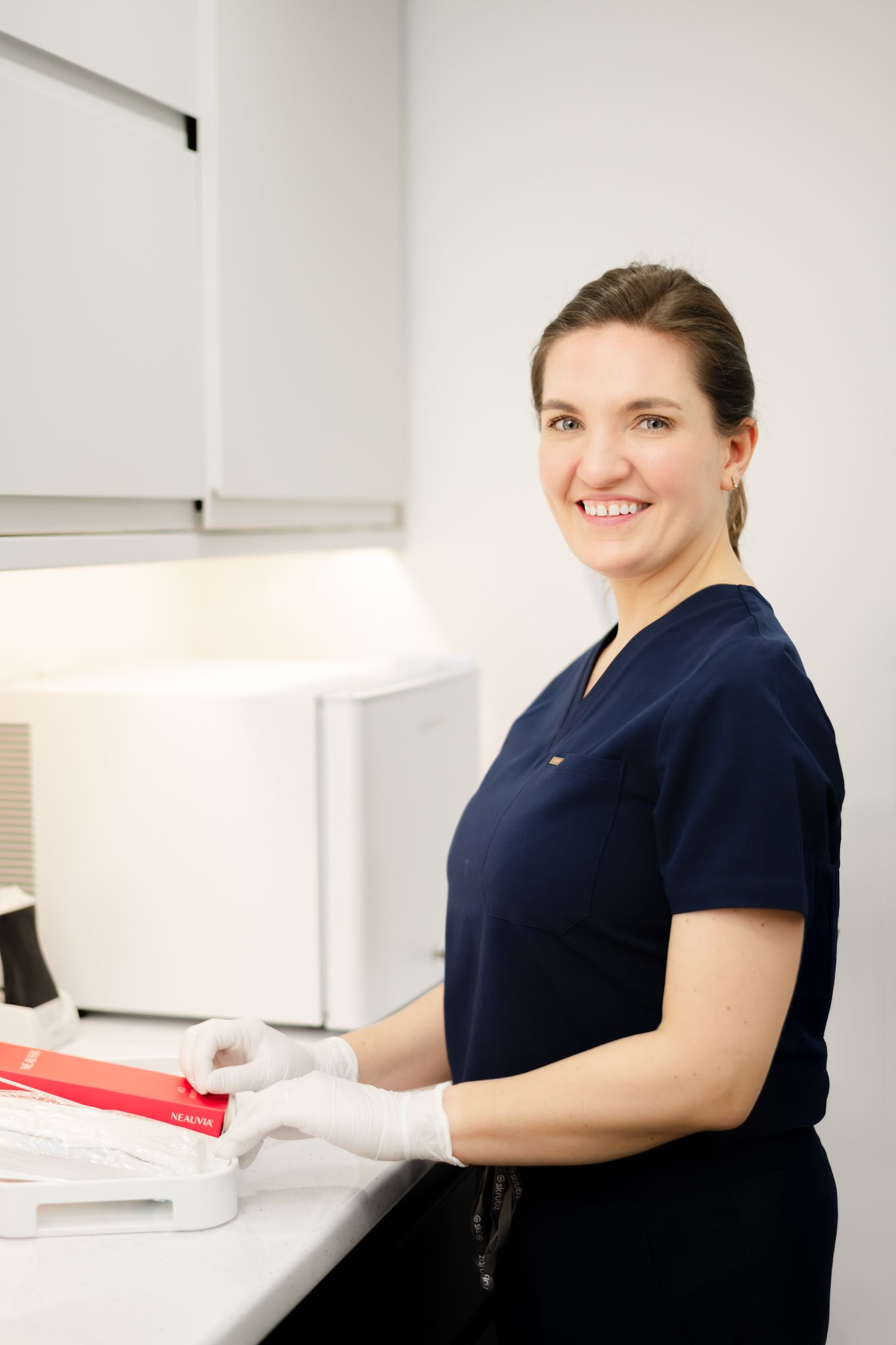 Medical professional in navy scrubs smiles while working at a white laboratory counter wearing protective gloves.