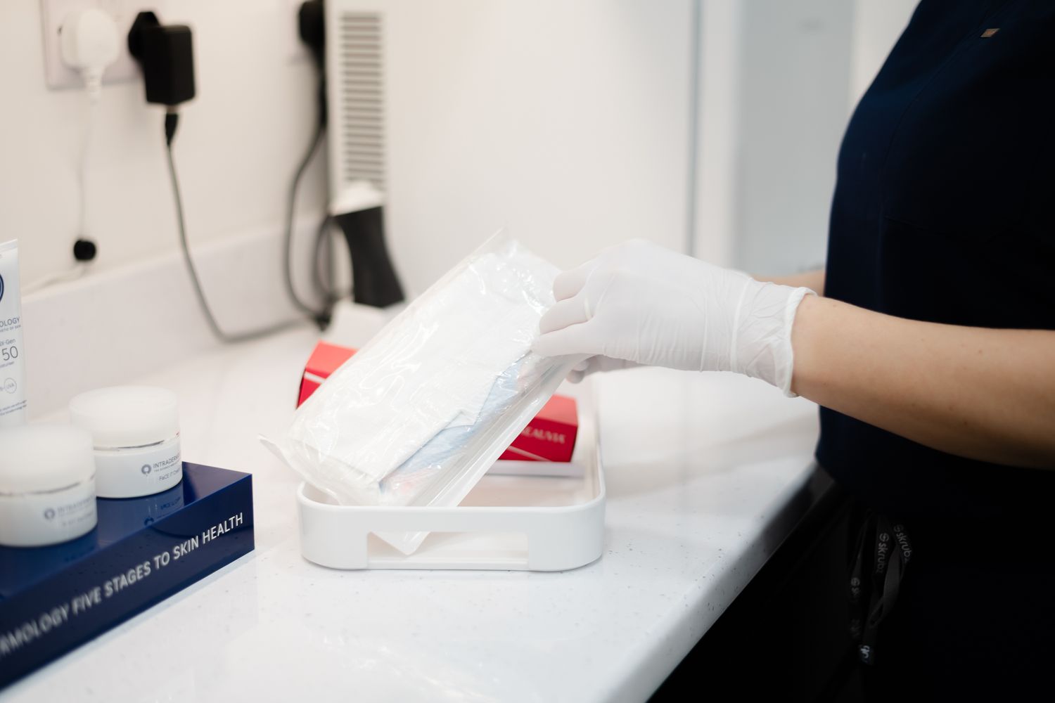 Gloved hands prepare beauty treatment supplies on white countertop with towels and skincare products.