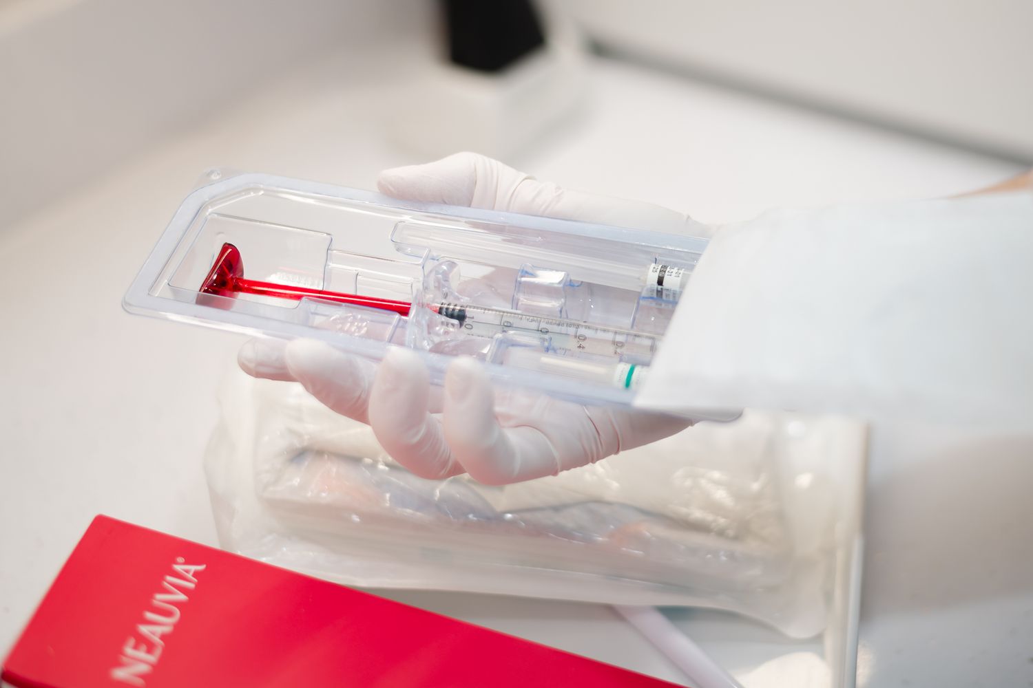 A clear medical syringe and red packaging materials sit on a bright white surface.