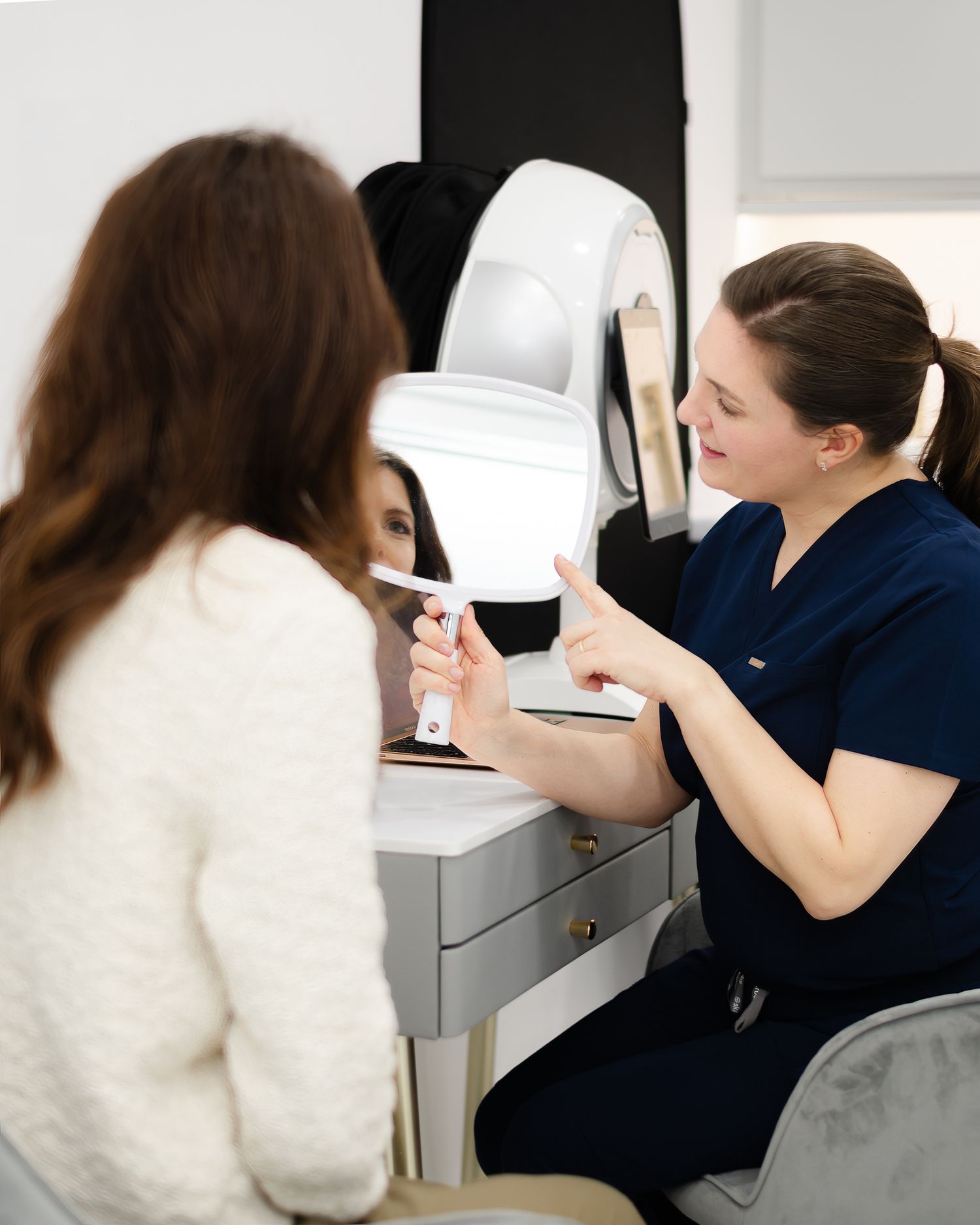 A healthcare provider in navy scrubs consults with a patient at a medical office desk.