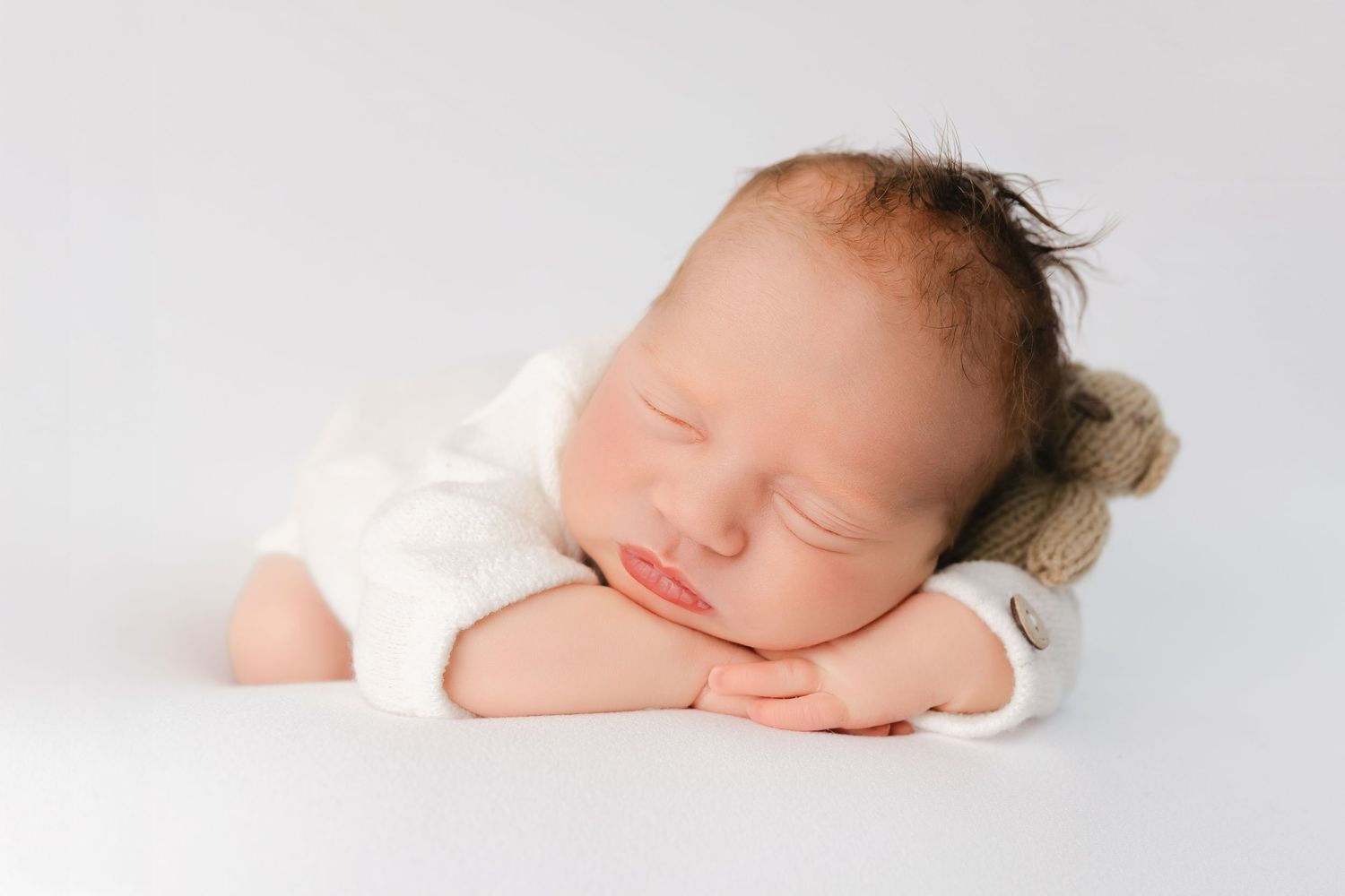 Peaceful sleeping newborn in white onesie rests with arms folded on light background.