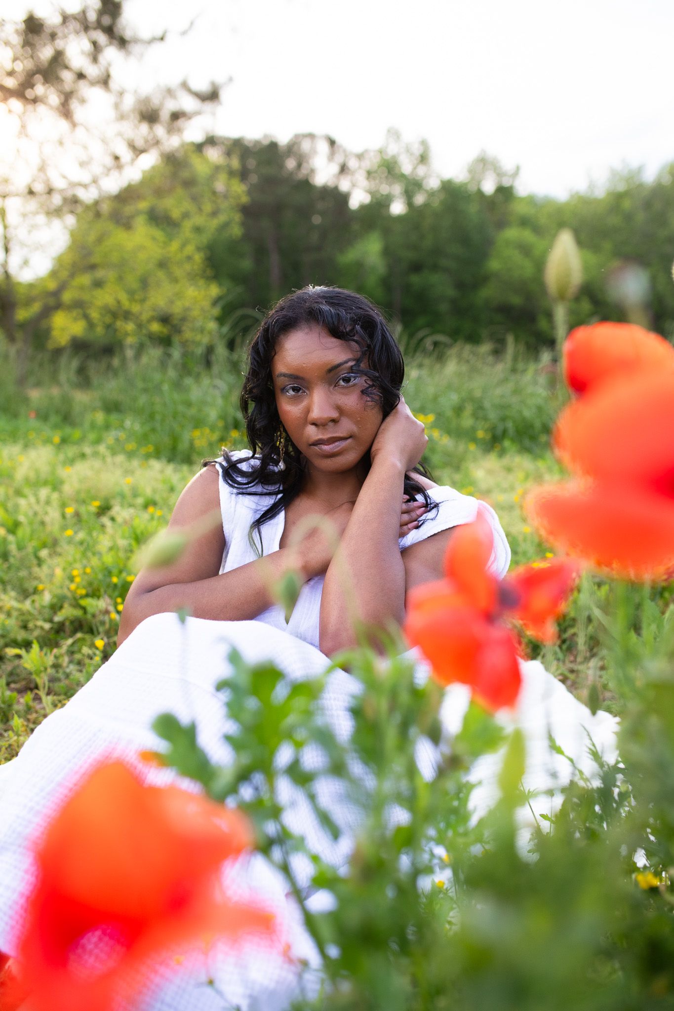 a Woman wearing white at a farm posing with red poppies framing the foreground and lush greenery in the background.