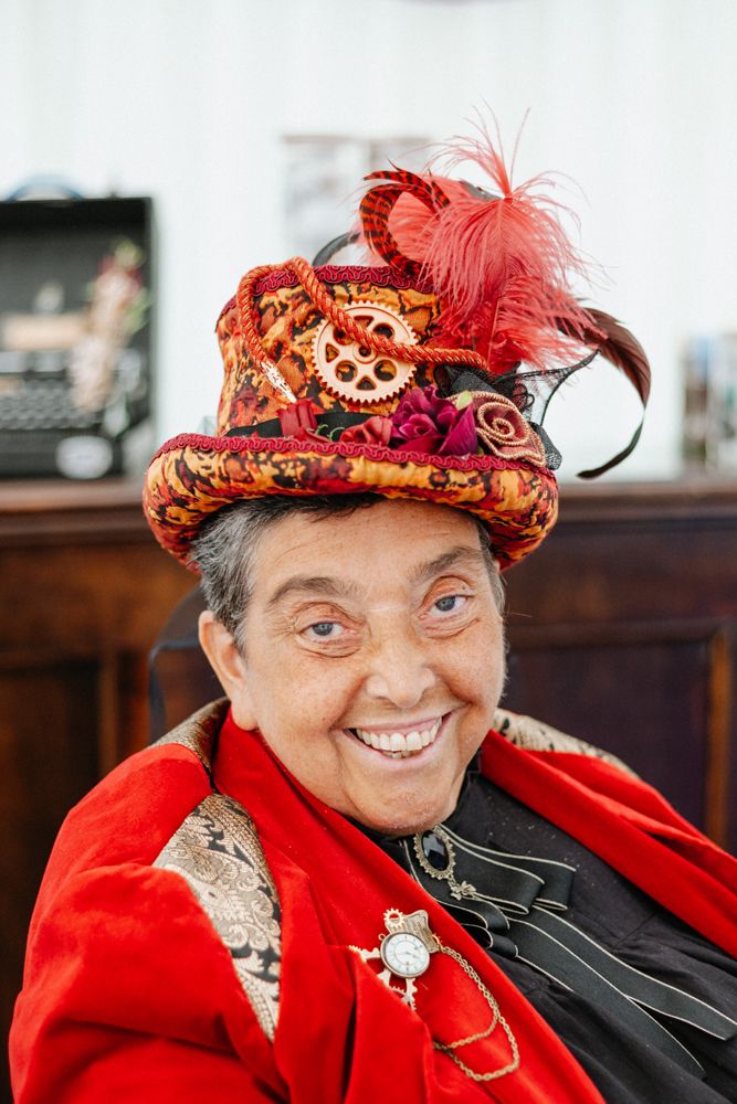 A steampunk-inspired top hat in red and gold with gears and feather decorations atop a bright red outfit.
