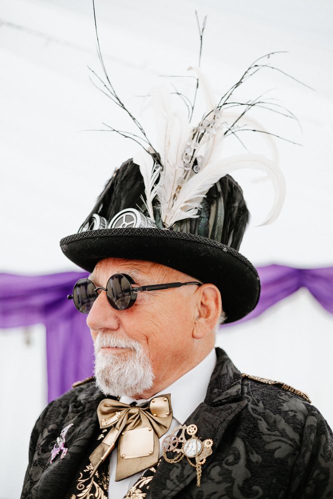A steampunk enthusiast wearing a black top hat adorned with white feathers and round sunglasses at a formal event.