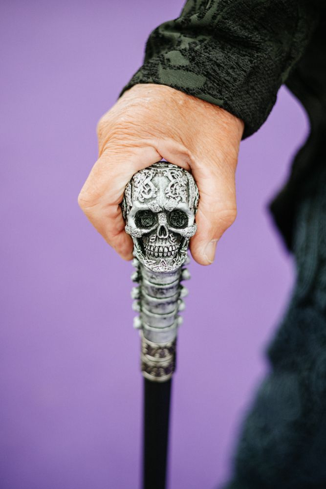 Hand holding an ornate walking cane with decorative silver skull handle against a purple background.