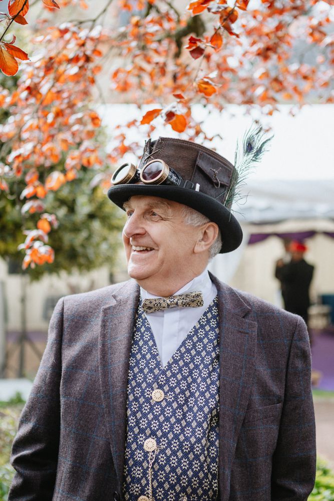 A person in steampunk attire wearing a top hat with goggles and patterned vest smiles beneath autumn foliage.