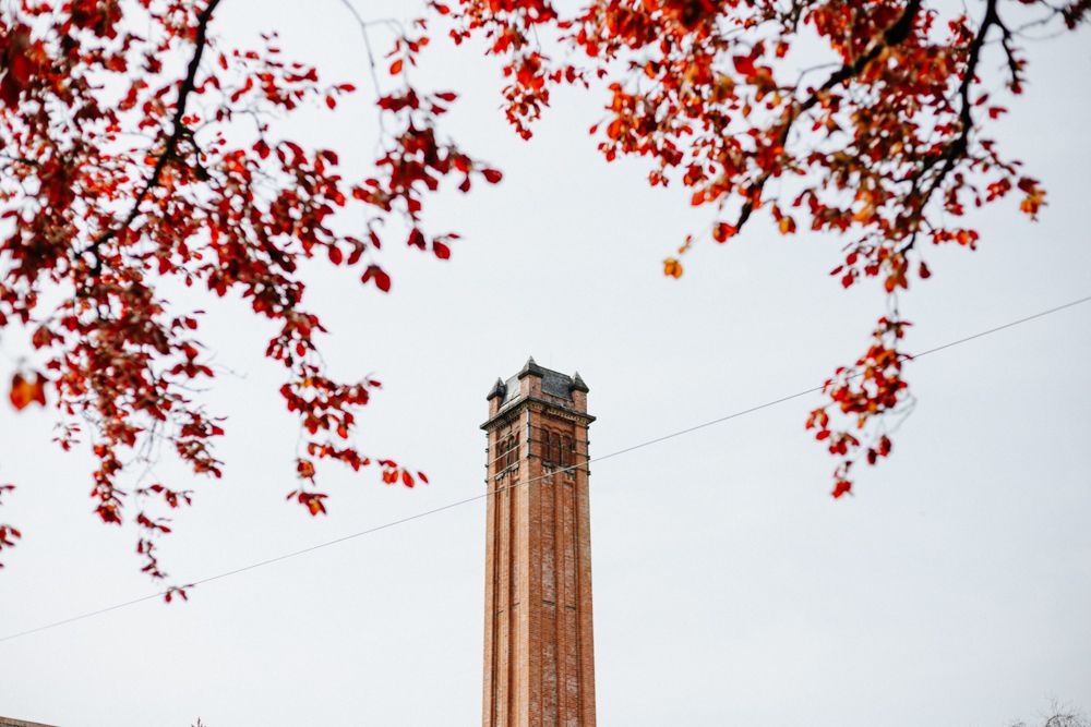 Tall brick chimney tower viewed through red autumn leaves against pale sky.