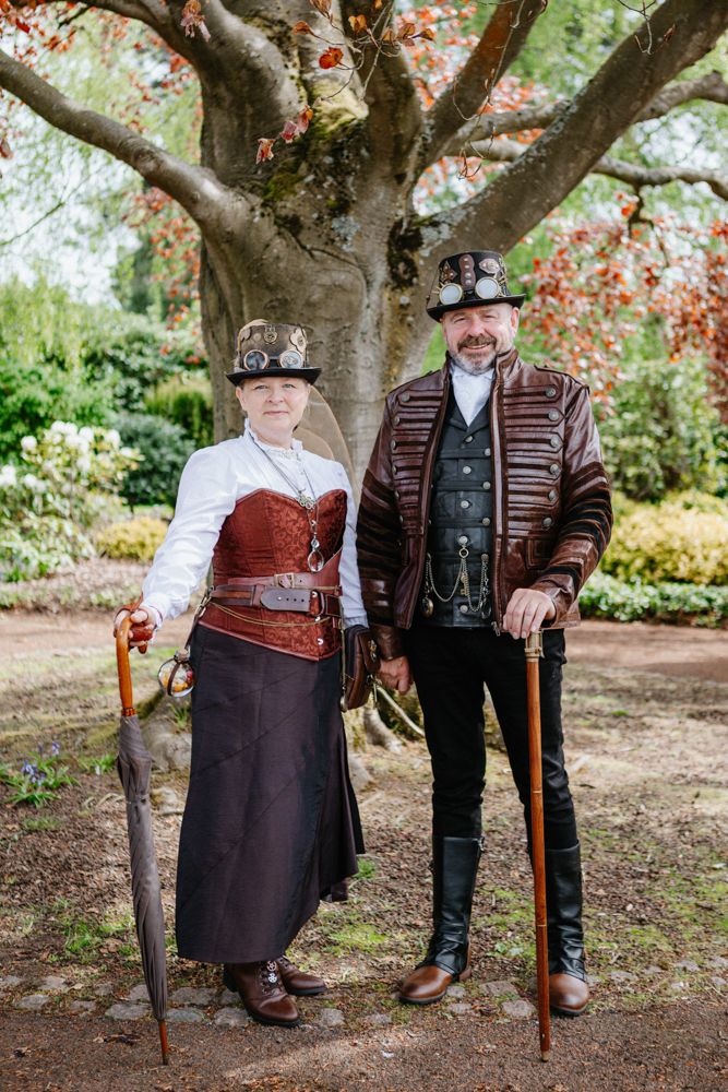 Two steampunk-styled figures in Victorian outfits with walking canes pose beneath a flowering tree in a garden setting.