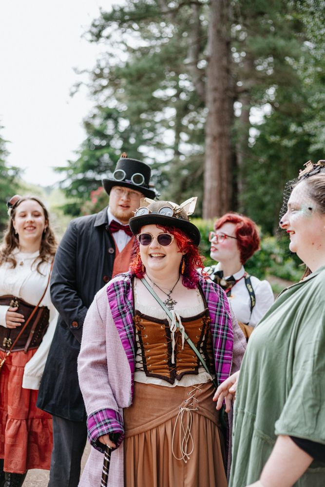 A group dressed in steampunk and Renaissance faire attire gathers outdoors in a wooded area, wearing period costumes.