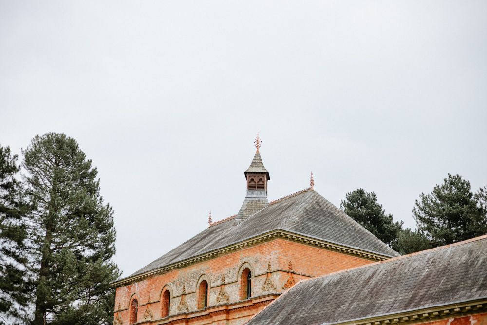 Historic orange brick building with ornate architectural details and cupola tower against cloudy sky surrounded by pine trees.