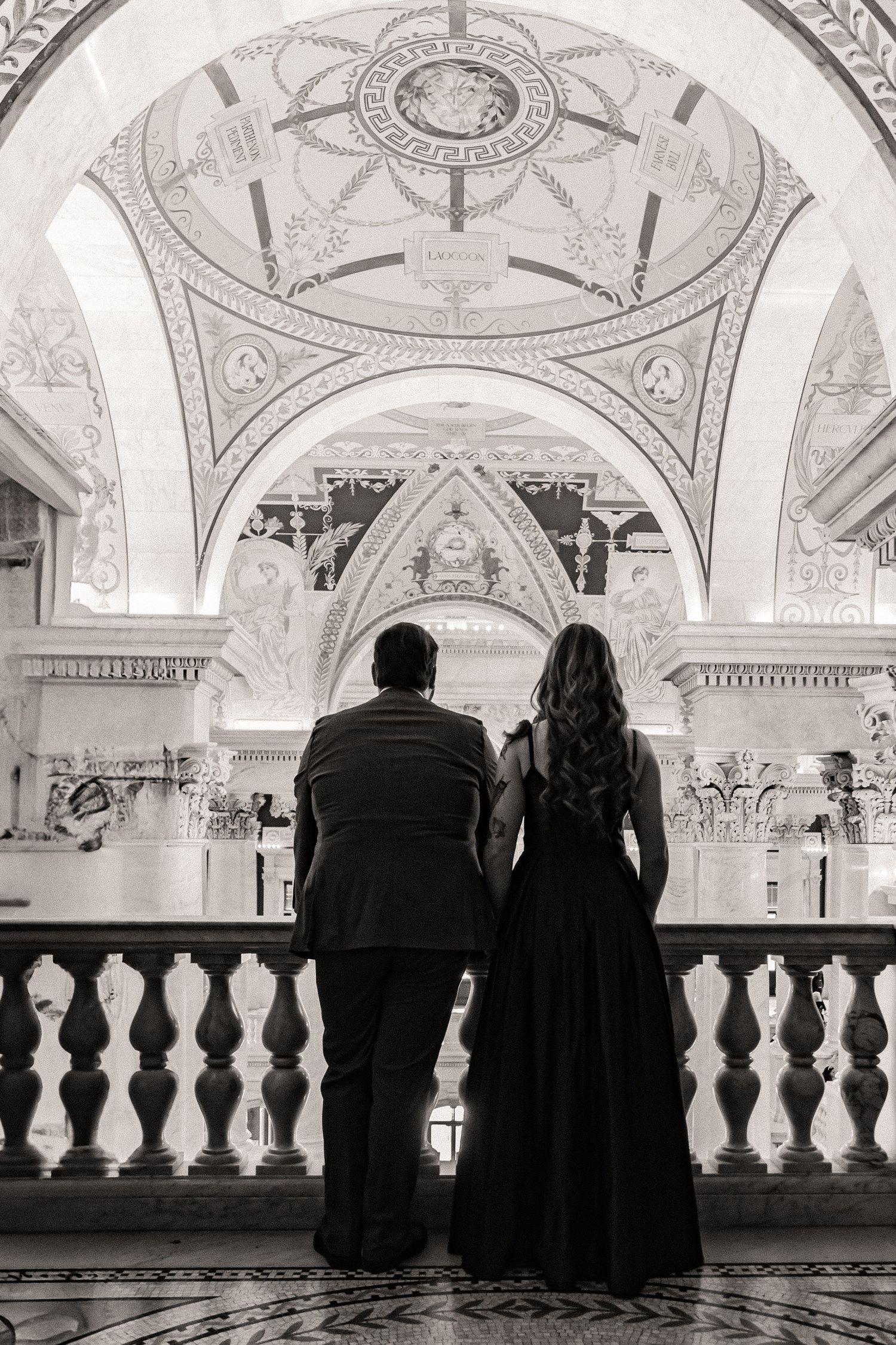 Two figures in formal dress stand at an ornate marble balustrade beneath an elegant domed ceiling with classical architecture.