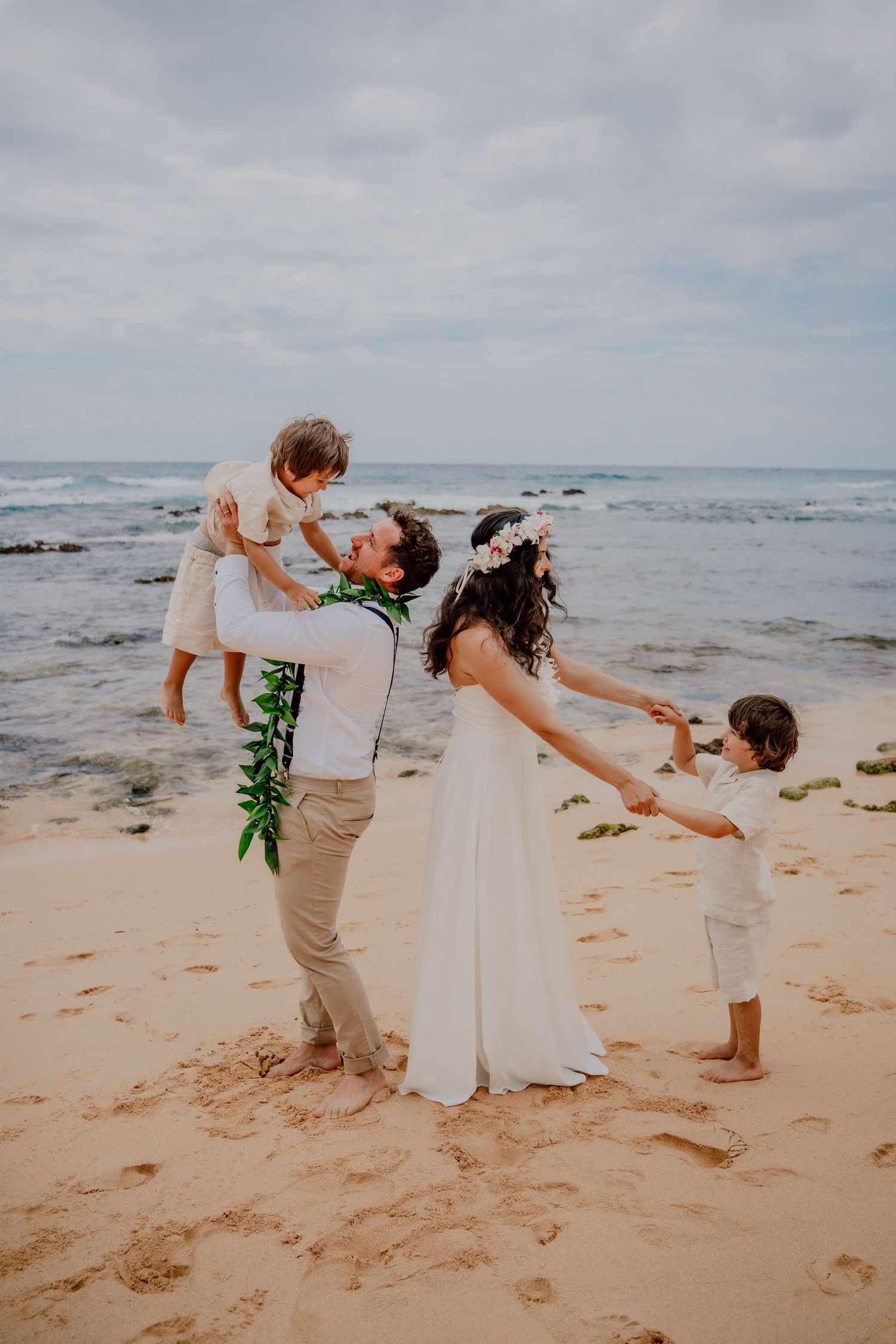 A family celebrates on a sandy beach in Hawaii with waves crashing in the background at sunset.