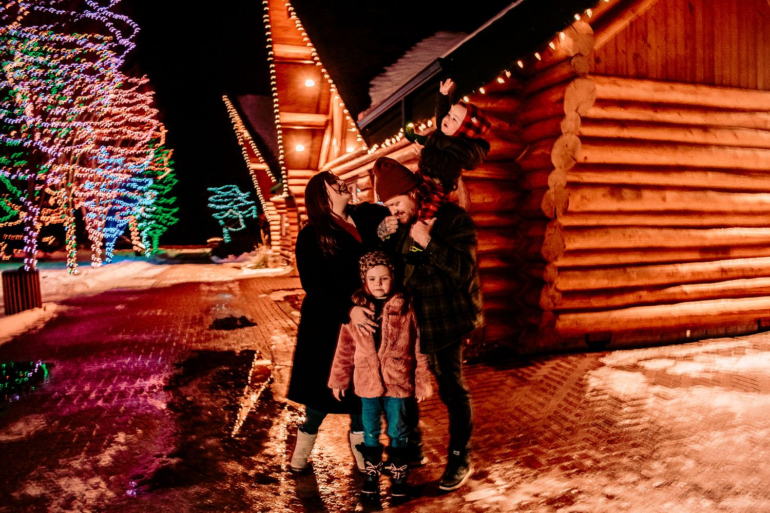 Group portrait outside log cabin with colorful holiday lights and snow on the ground.