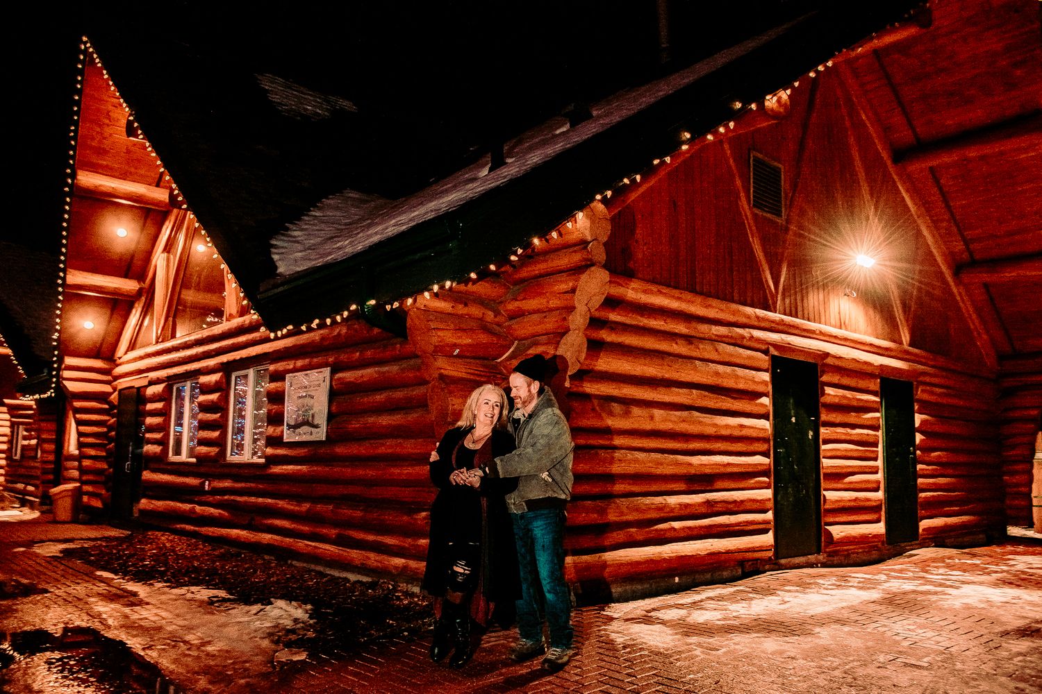 A romantic nighttime scene outside illuminated red log cabins with string lights creating a cozy winter atmosphere.