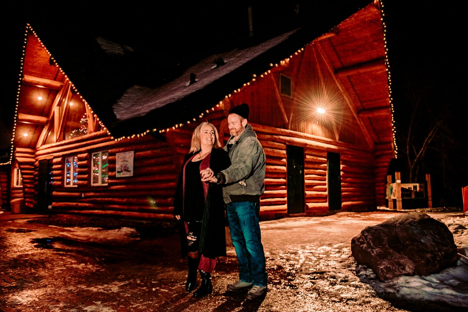 Couple embracing in front of warmly lit rustic log cabins decorated with Christmas lights on a snowy evening.