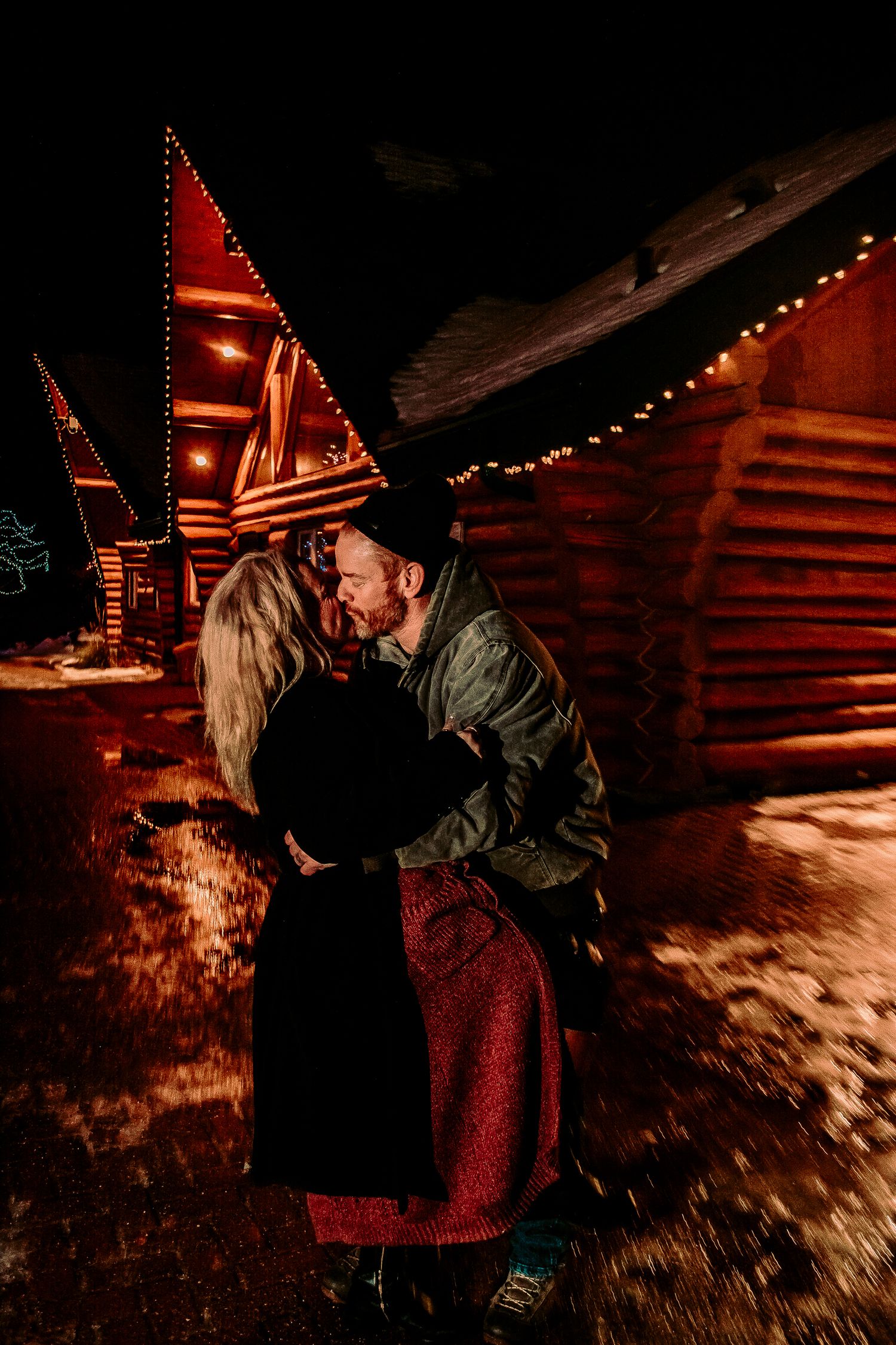 Romantic embrace outside snow-covered log cabins illuminated by warm red lighting and string lights.