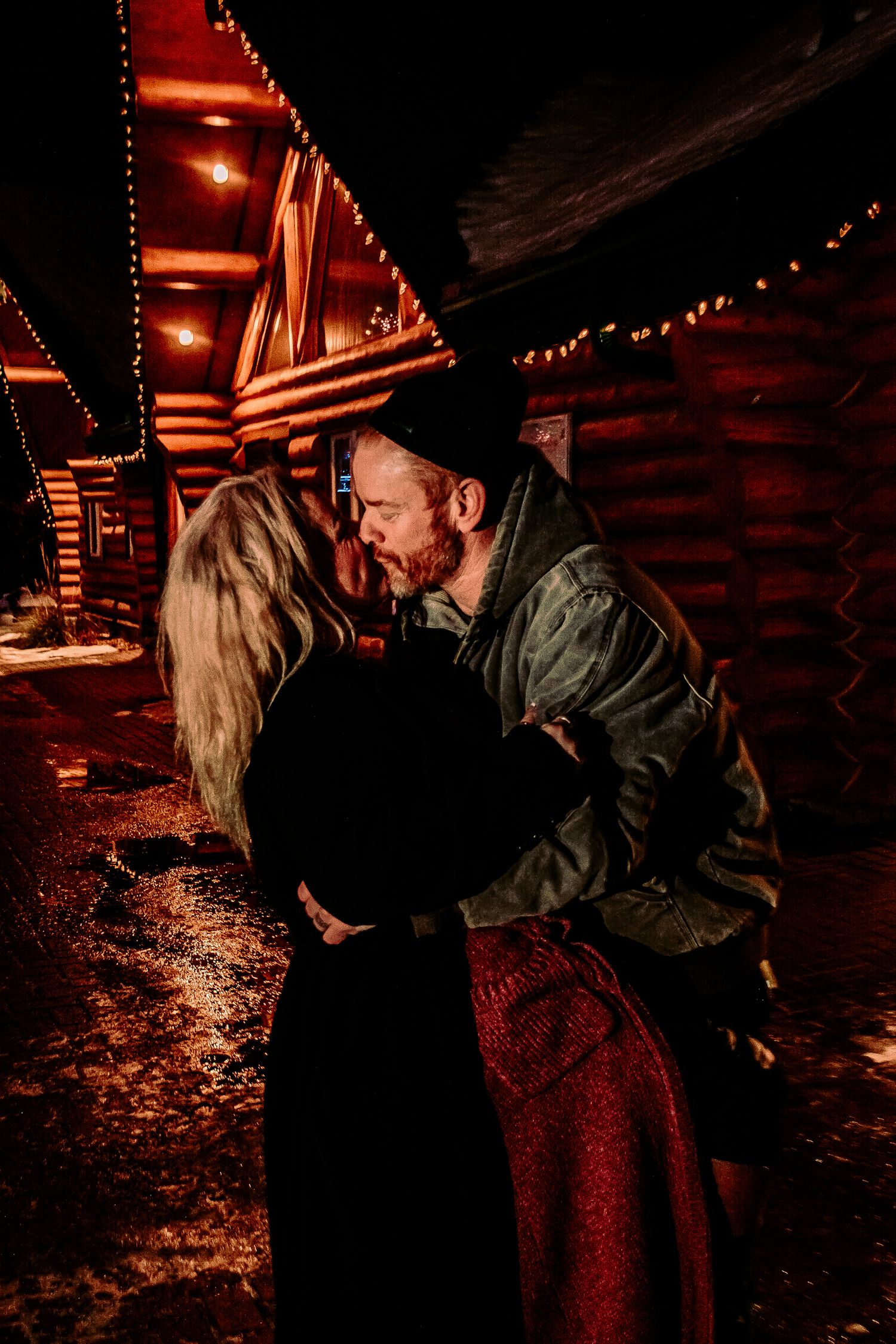 Couple sharing an intimate moment against the backdrop of illuminated log cabins on a winter night.