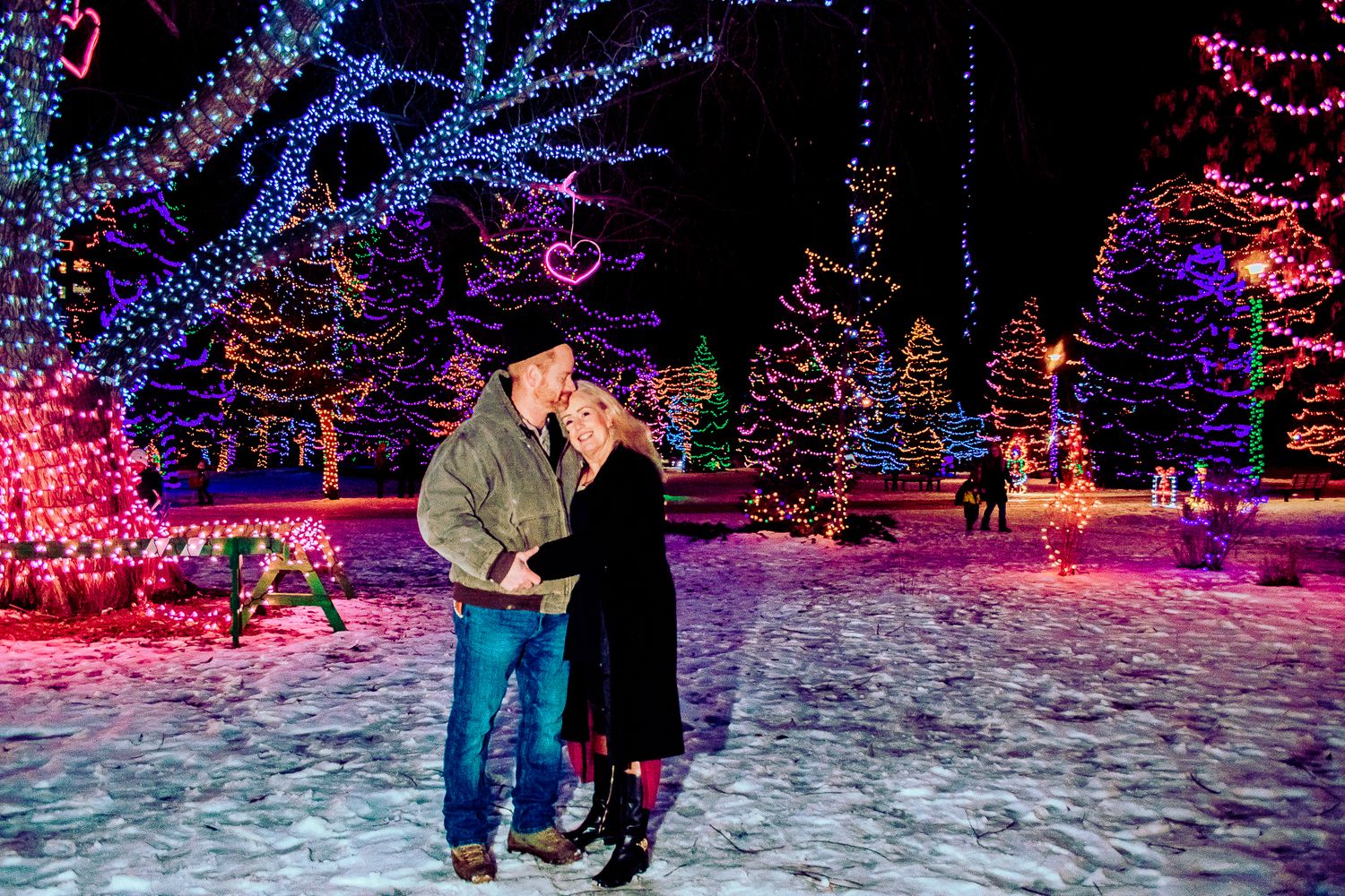 Couple surrounded by colorful Christmas lights and decorated trees in a magical winter wonderland setting.