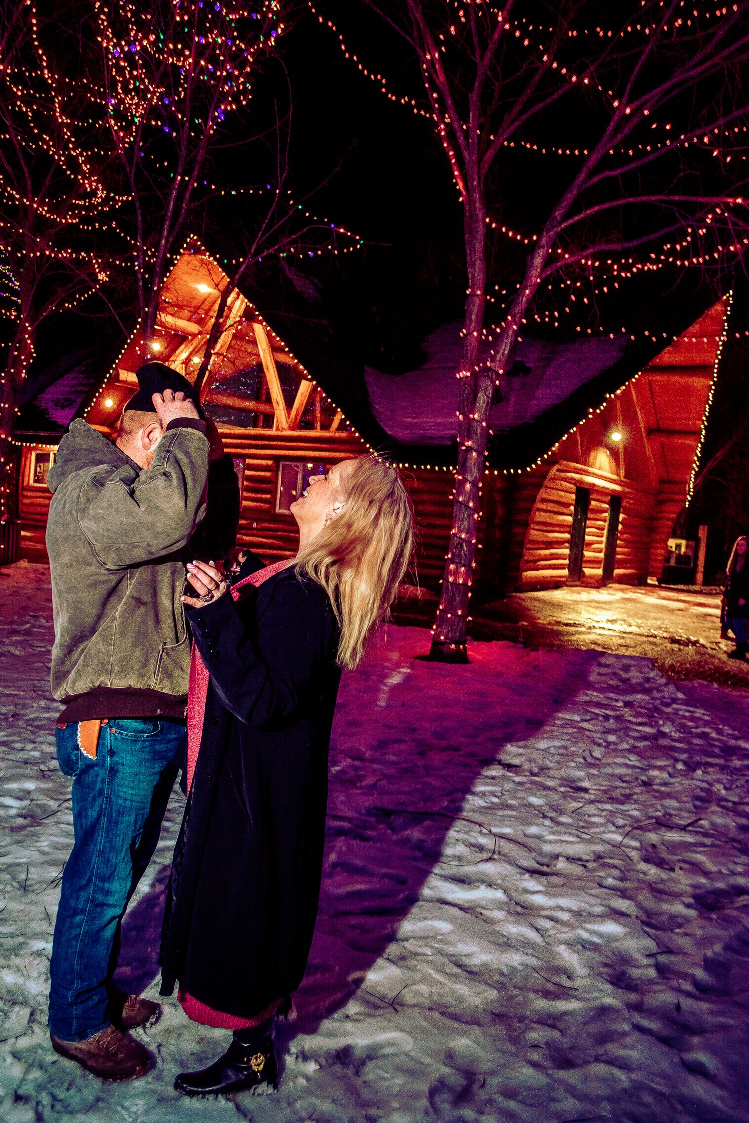 Romantic winter night scene with couple by log cabin surrounded by trees wrapped in purple holiday lights.