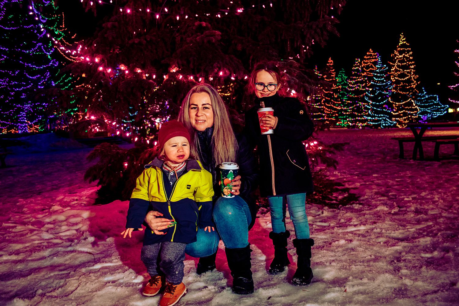 Family posing in snow beneath pink holiday lights with illuminated Christmas trees behind them.