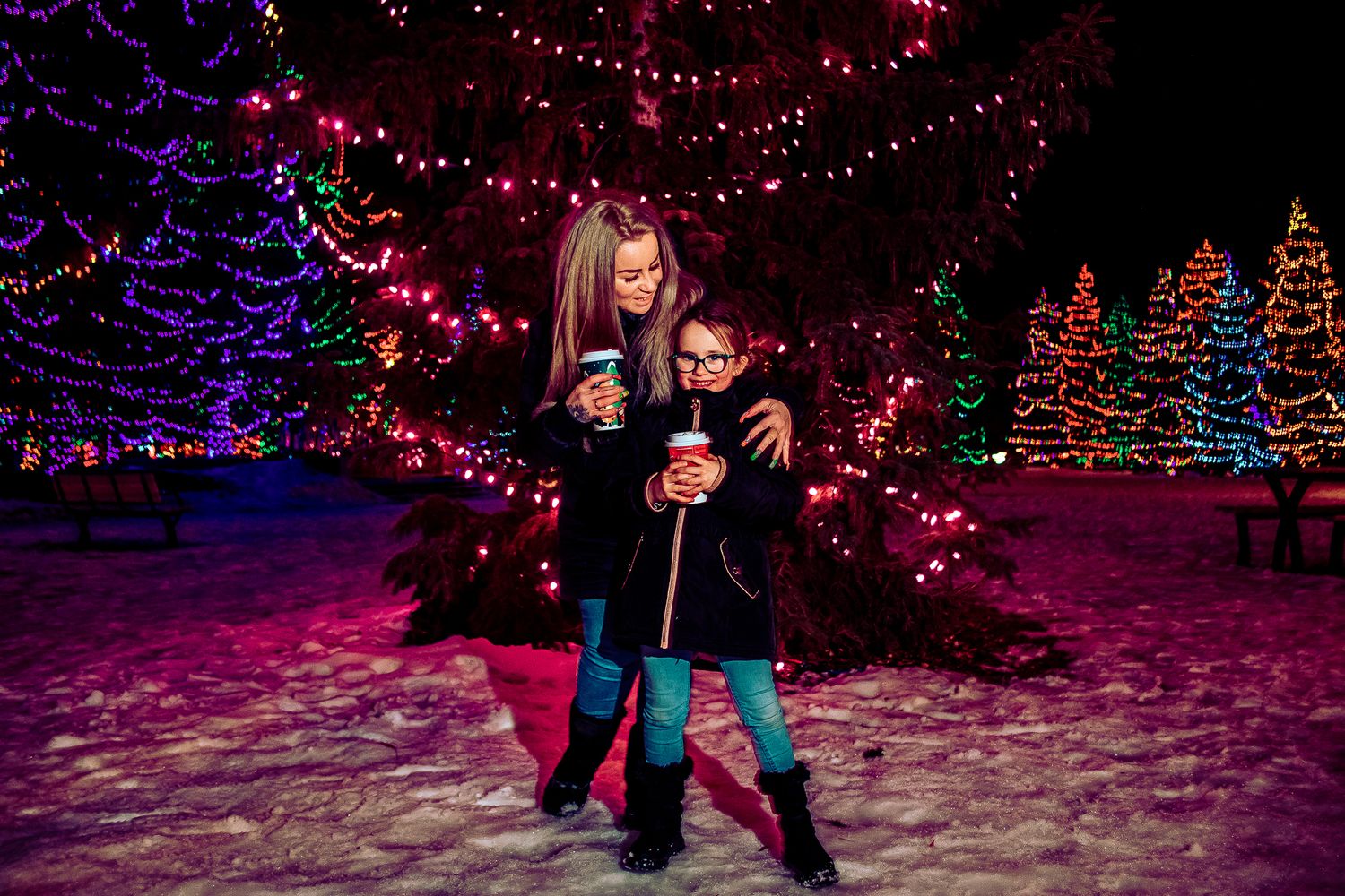 Two people pose in front of illuminated Christmas trees decorated with festive red and purple lights in the snow.