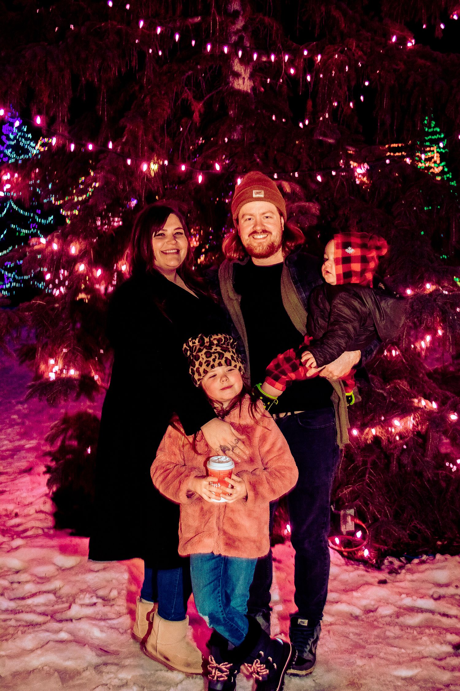 A family poses together amid colorfully lit trees and holiday decorations in a snowy winter scene.