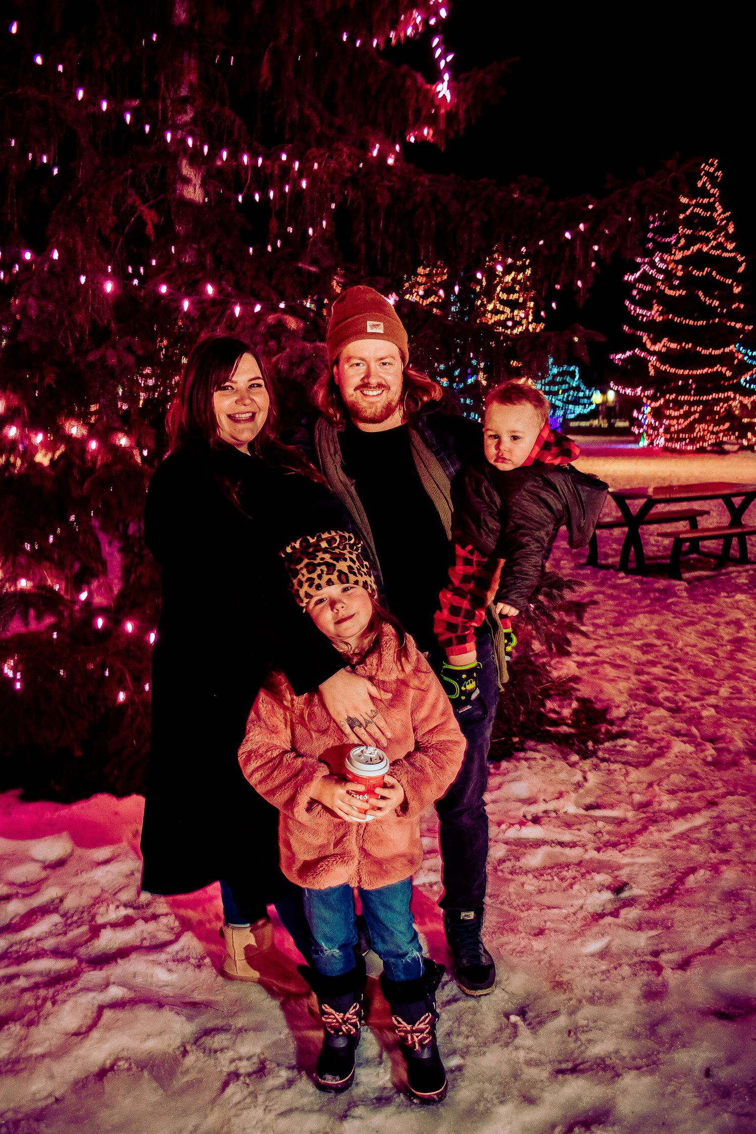 A family group stands together against a backdrop of glowing red Christmas lights and illuminated trees.