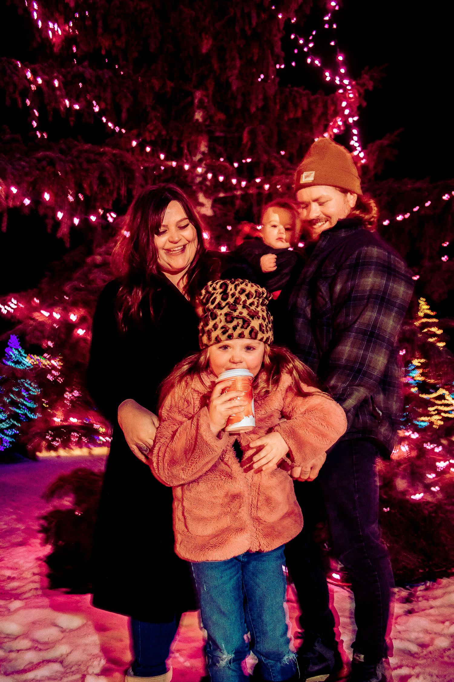 A family portrait captured against a Christmas tree with bright pink lights and neon trees in the background.