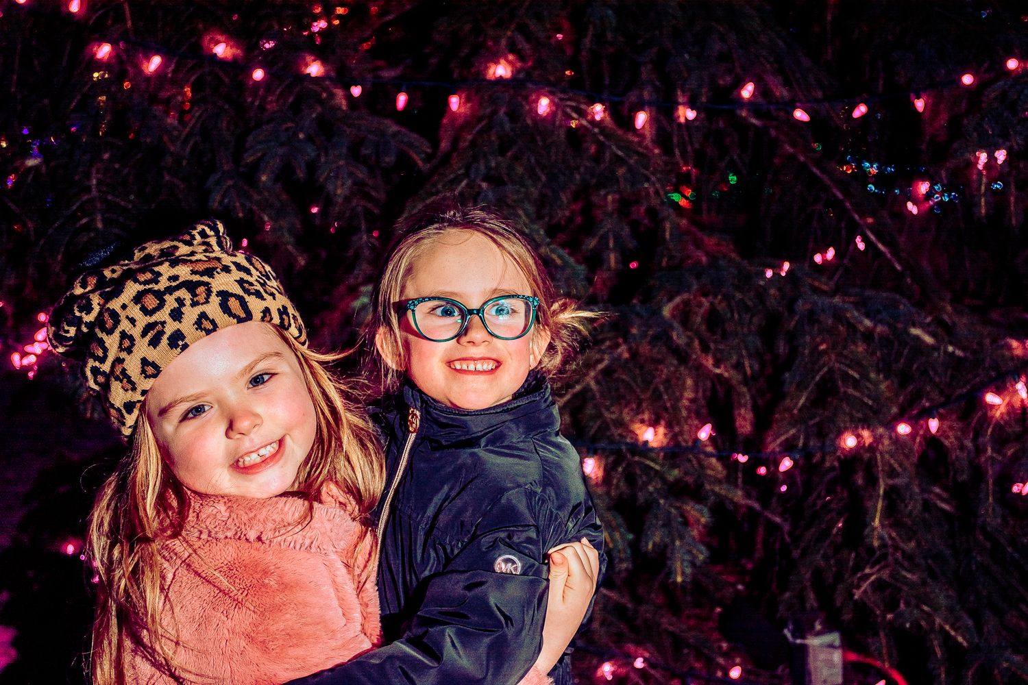 Two bundled up children pose together under festive pink holiday lights.