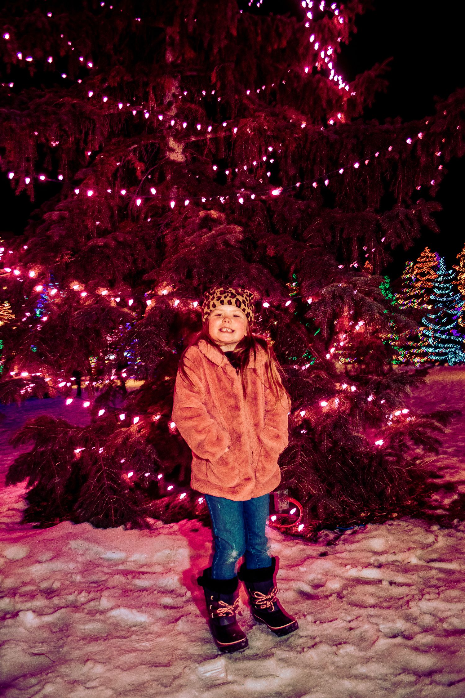 Full-length winter portrait in front of a large tree decorated with pink holiday lights.