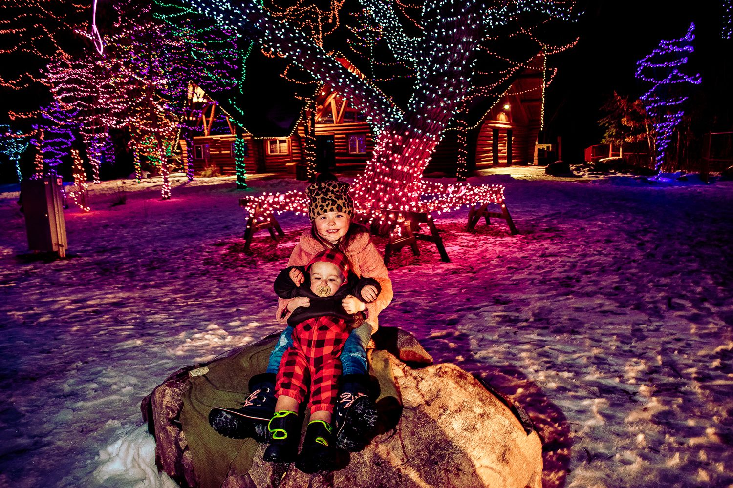 People huddled together on a snowy rock surrounded by colorfully lit holiday decorations and houses.
