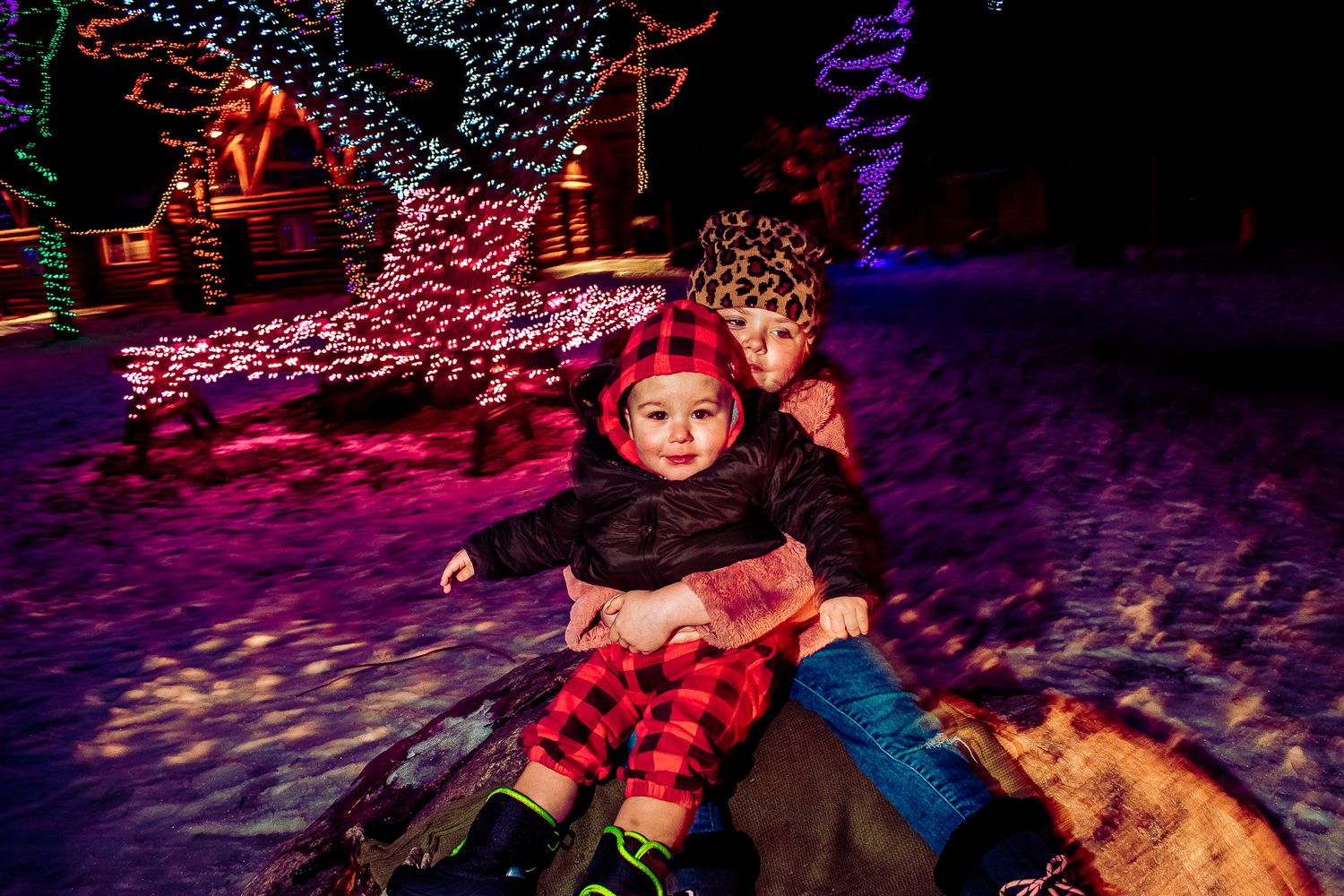 People sitting playfully on a rock with colorful Christmas lights illuminating the snowy scene.