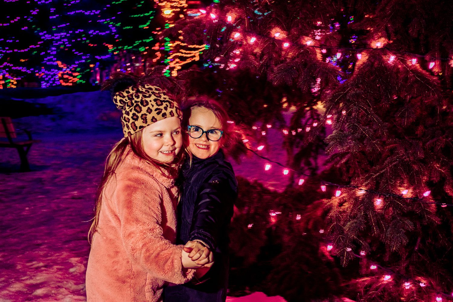 Two children embrace while surrounded by twinkling holiday lights in the snow.
