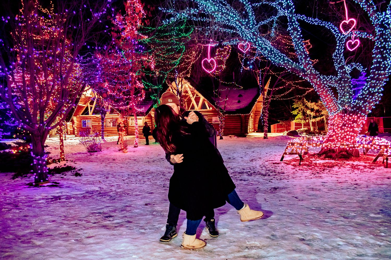 Two people dancing in the snow surrounded by colorful holiday lights and decorated buildings.