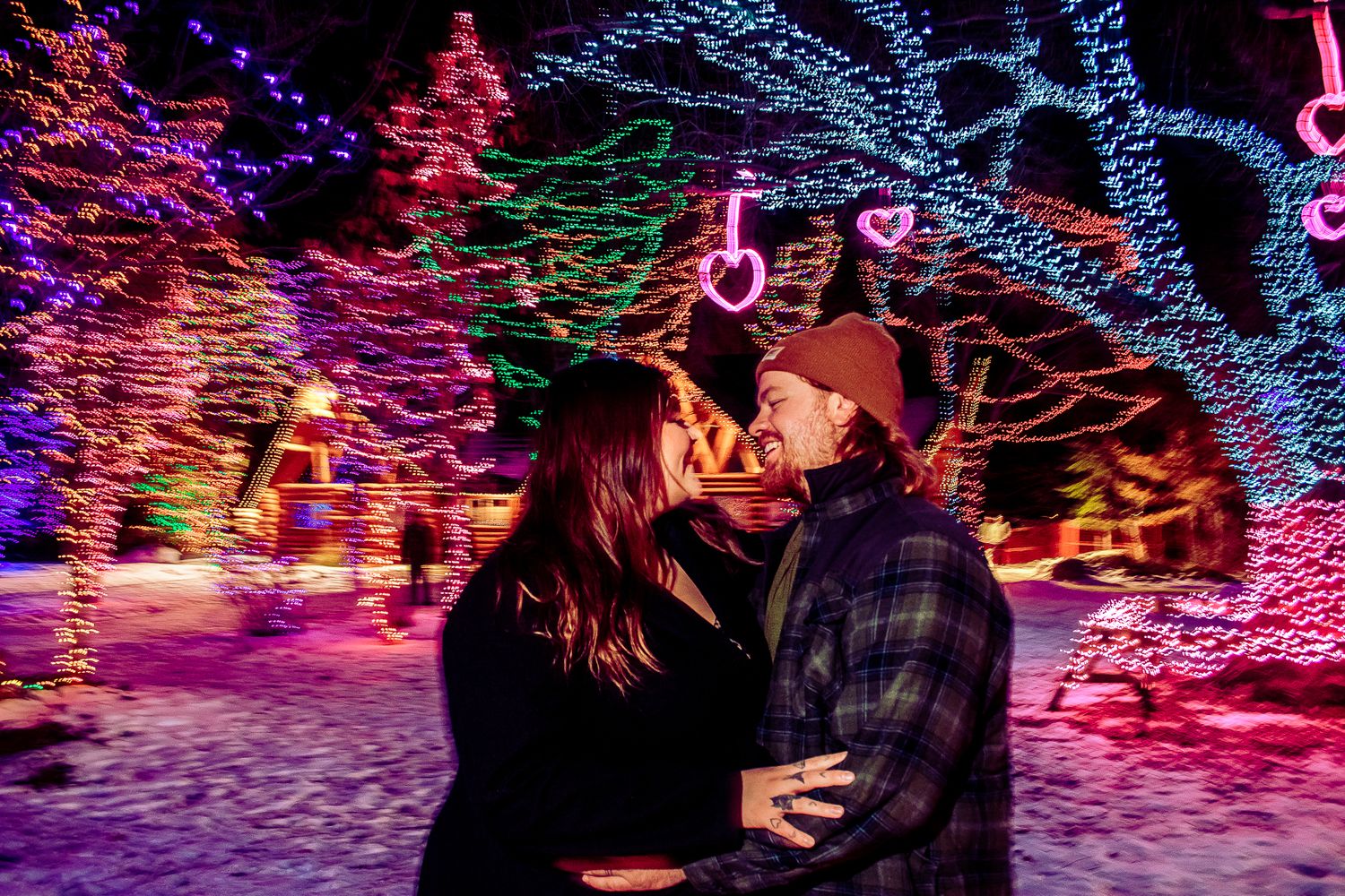 A romantic moment captured under festive holiday lights with snow-covered scenery in the background.