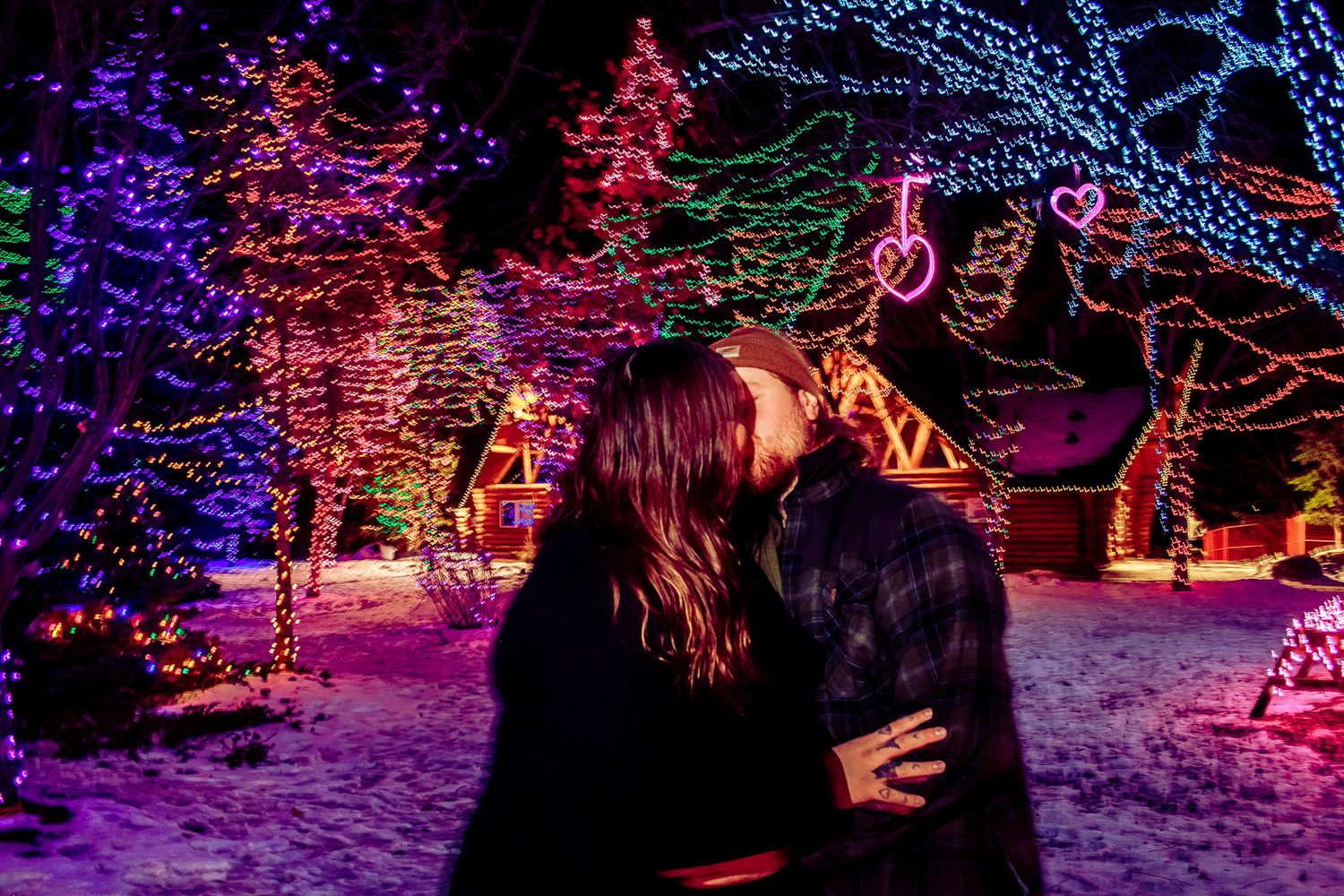 Two people embracing under colorful Christmas lights in a snowy winter wonderland display.