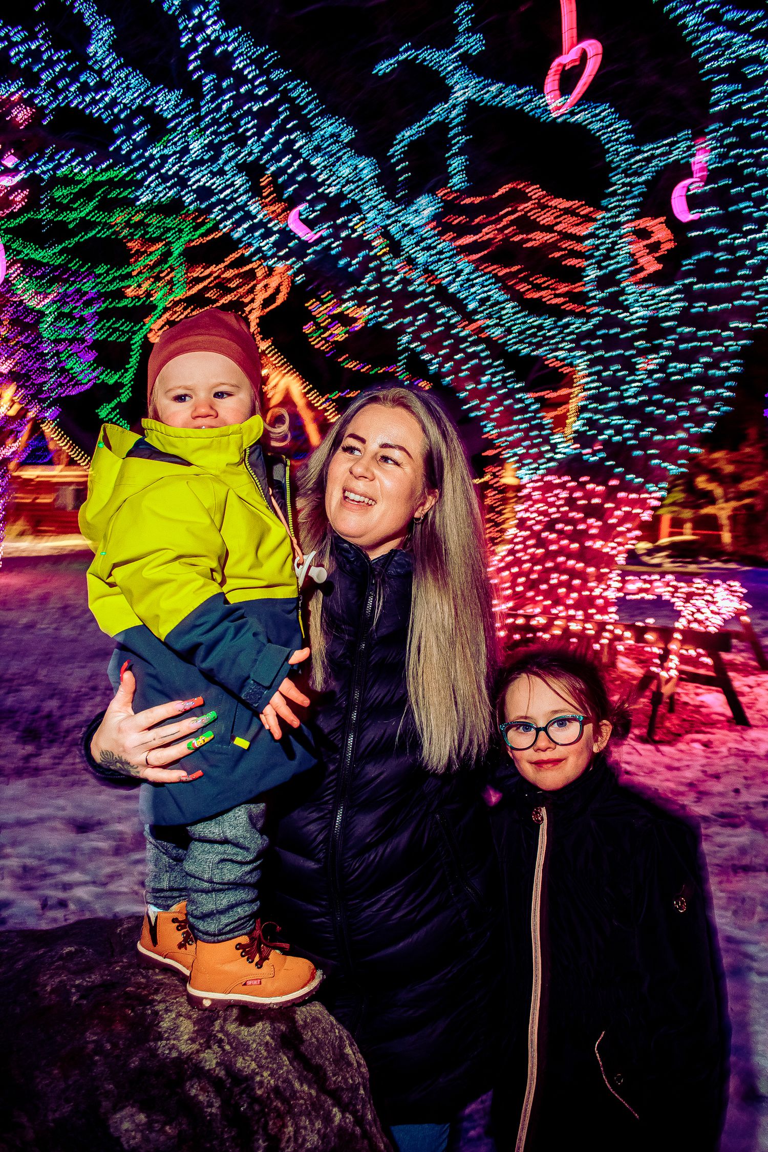 Family enjoying holiday lights display while posing on a rock in winter setting.