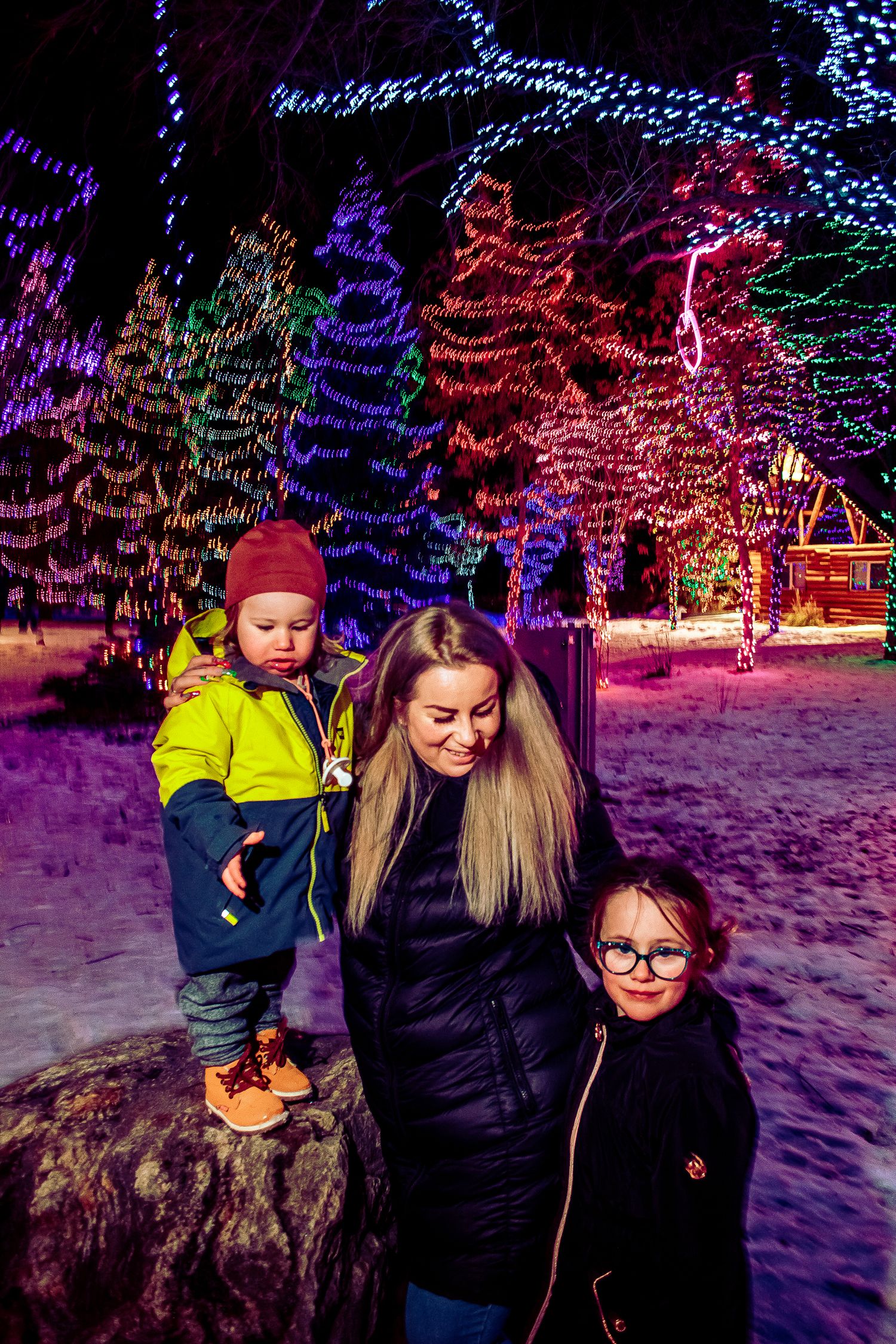 Family members posing together among colorfully lit Christmas trees in snowy evening scene.