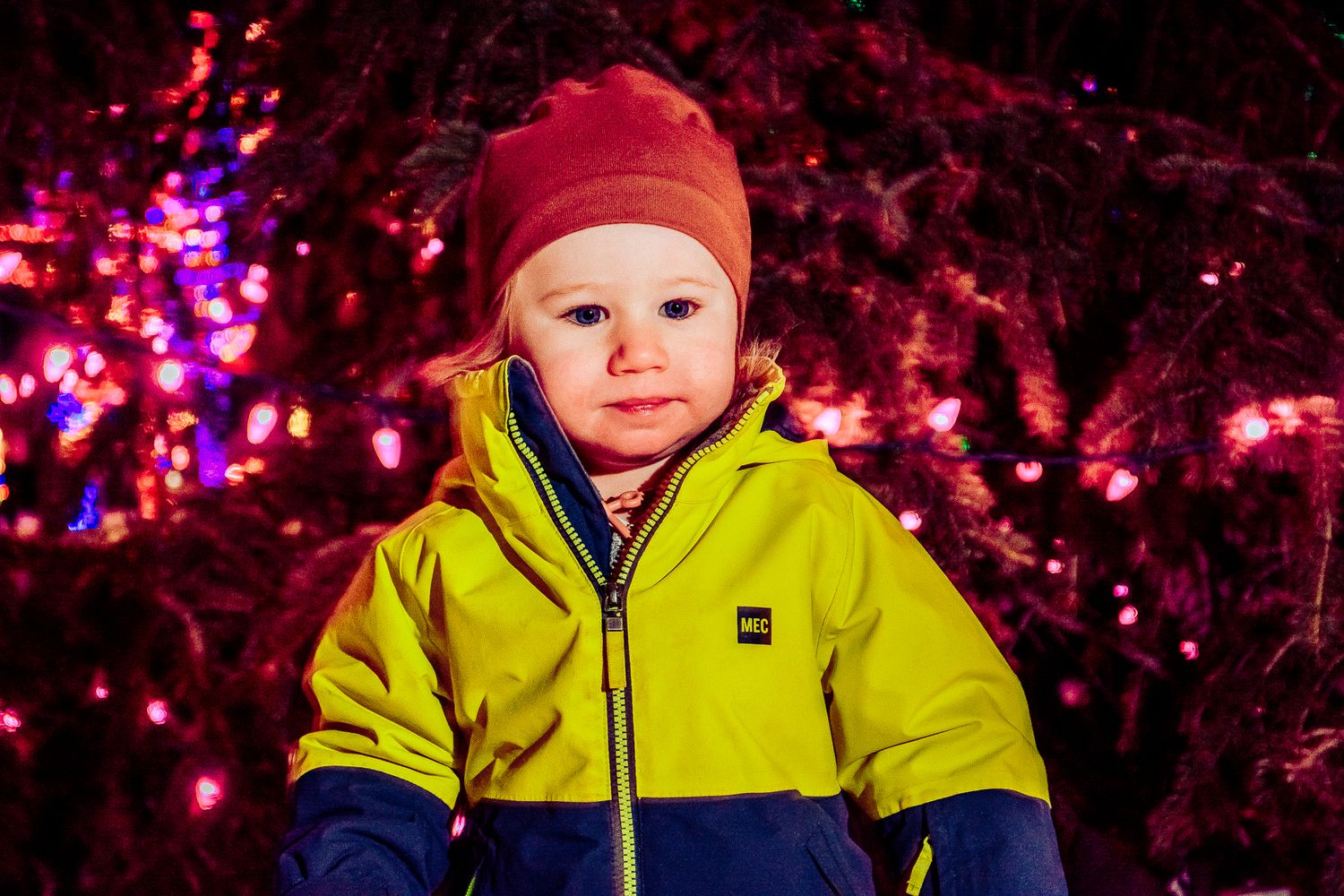 A child in bright winter clothing poses in front of illuminated holiday decorations.