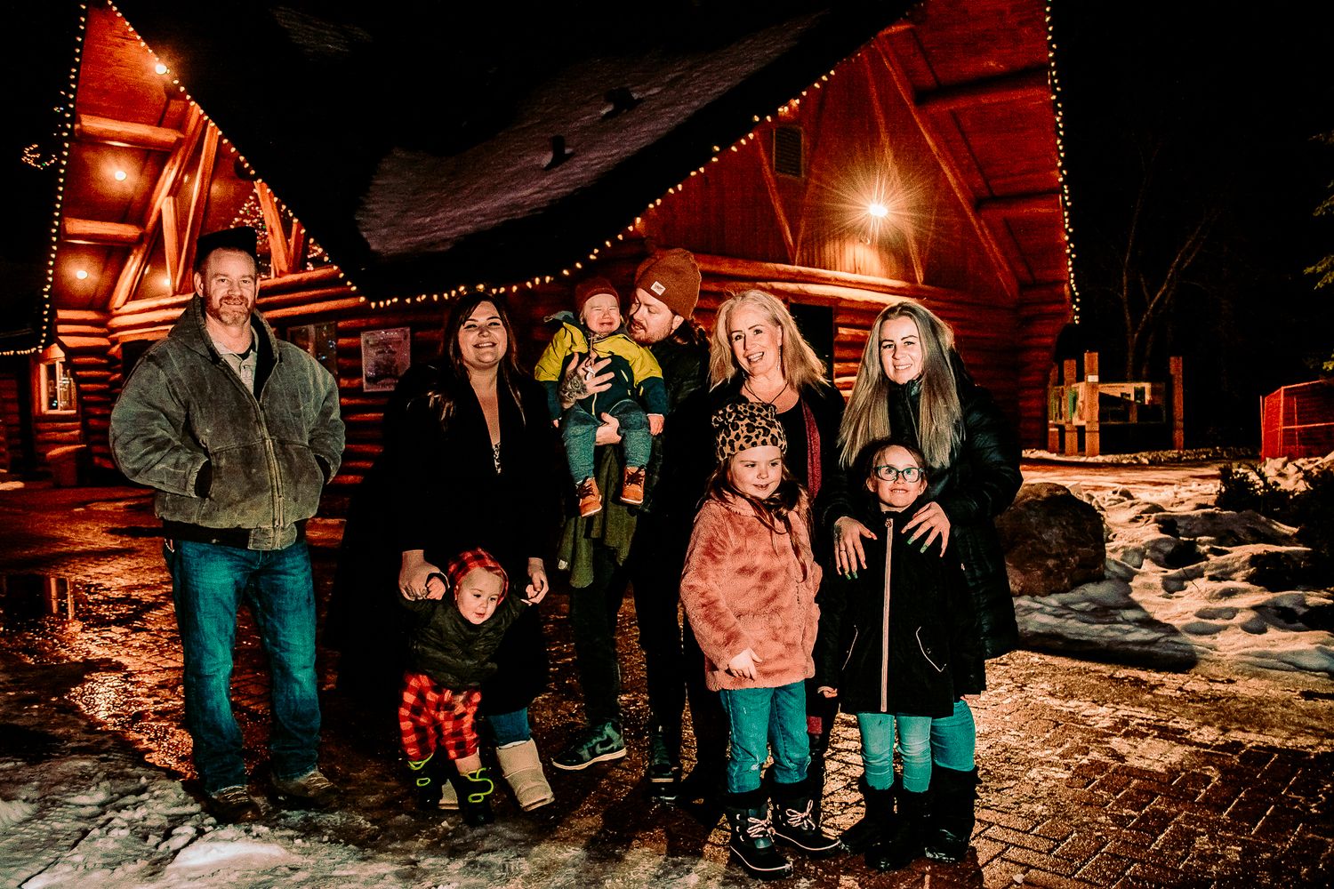 A group poses together outside a cozy log cabin decorated with Christmas lights on a snowy winter evening.