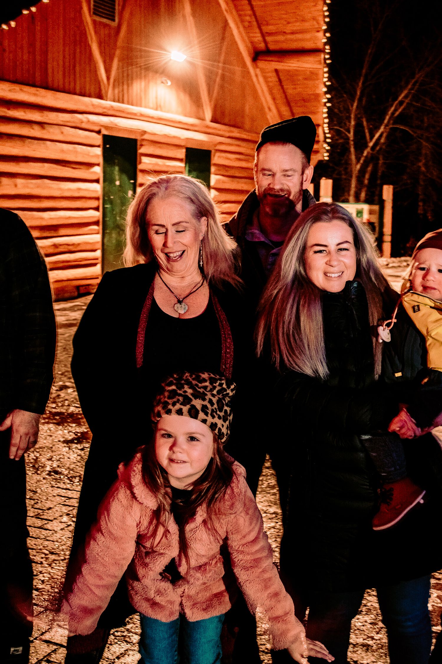 A joyful gathering outside a rustic log cabin illuminated by string lights on a winter night.