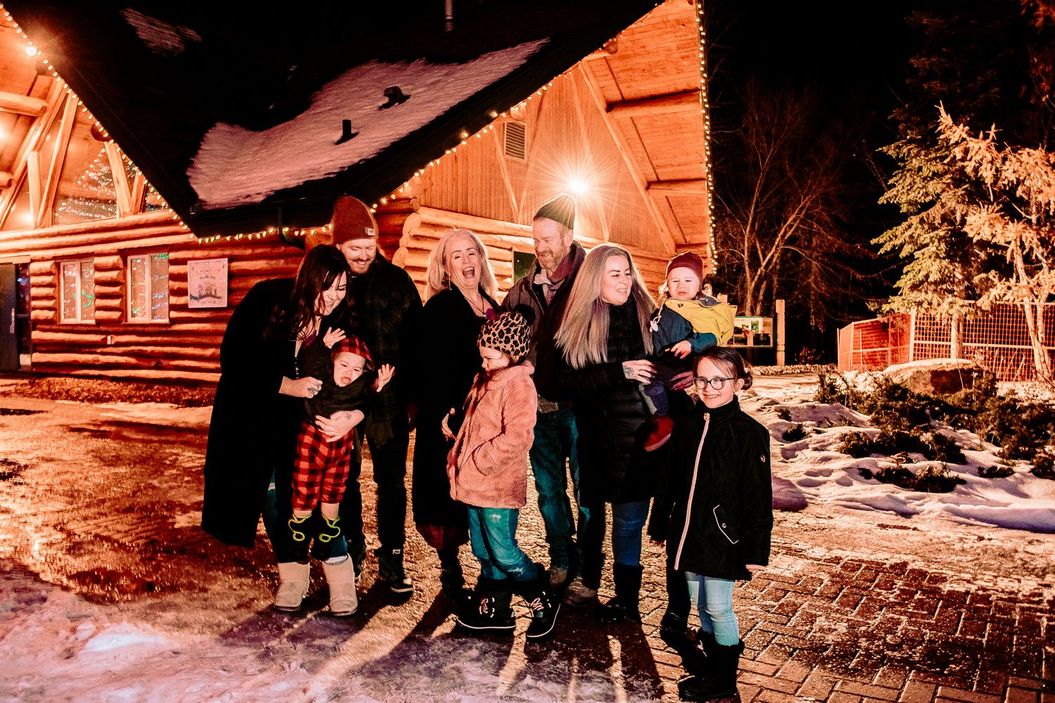 Group stands outside a snow-covered log cabin with festive holiday lights.