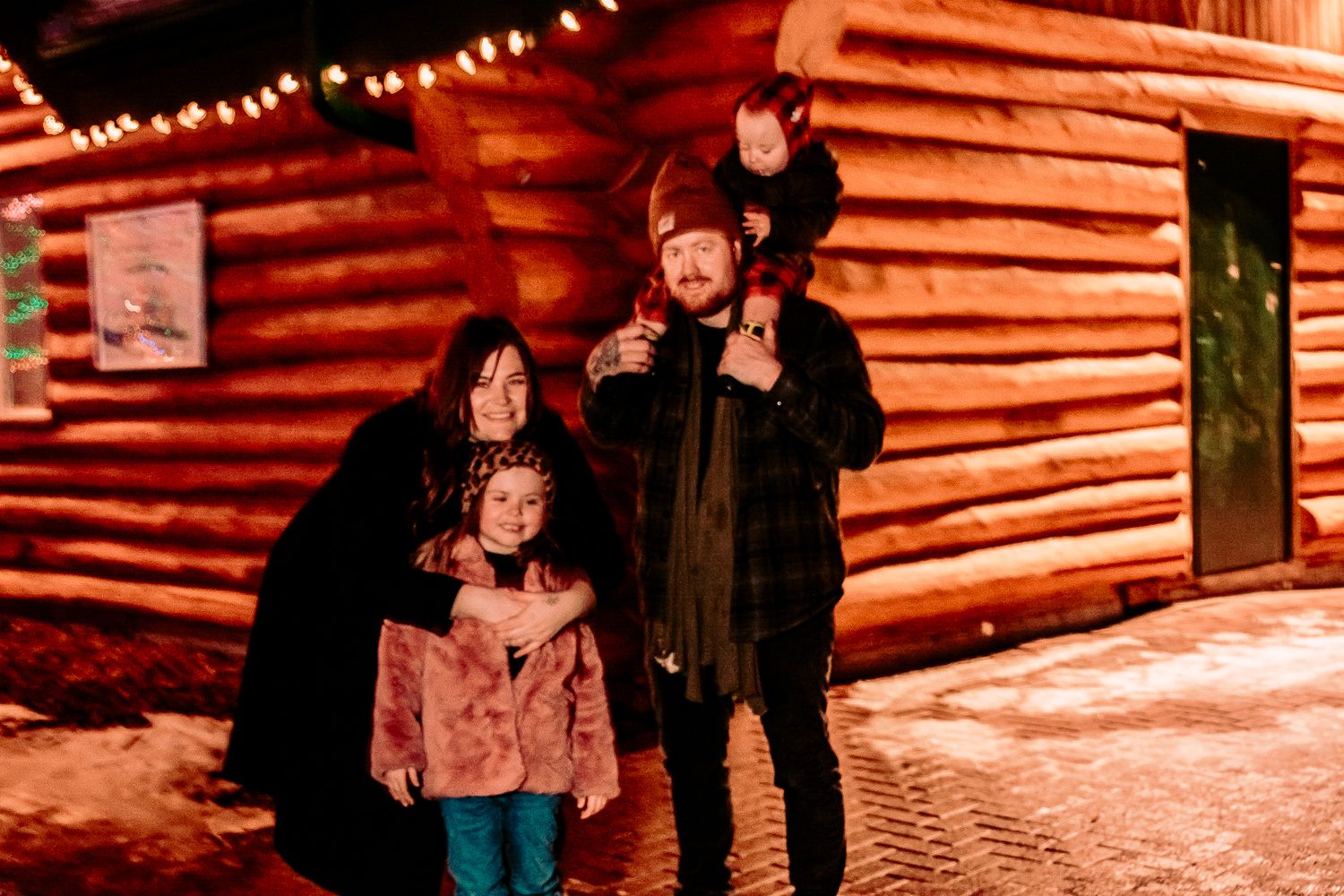 Family poses together at night outside an illuminated wooden cabin in winter.