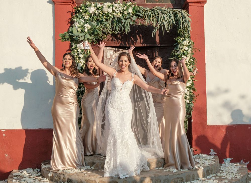 Celebratory wedding party in champagne-colored dresses poses beneath rustic archway adorned with greenery.