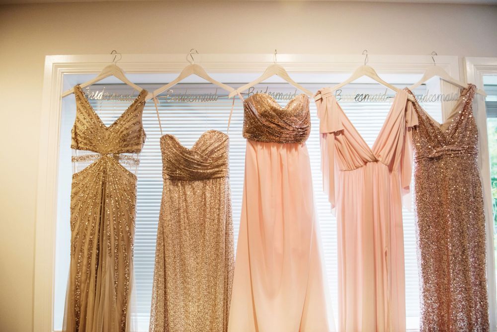 Five elegant bridesmaid dresses in blush pink and gold tones hanging in a row on white hangers against a light wall.