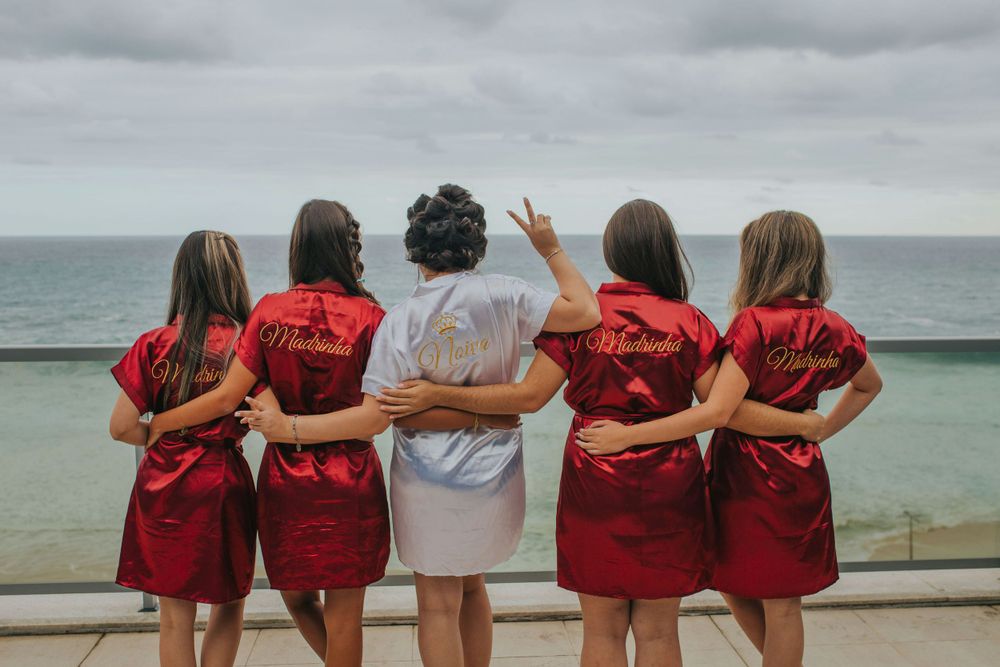 Group of friends in red and gray robes stand embracing while looking out at the ocean under cloudy skies.