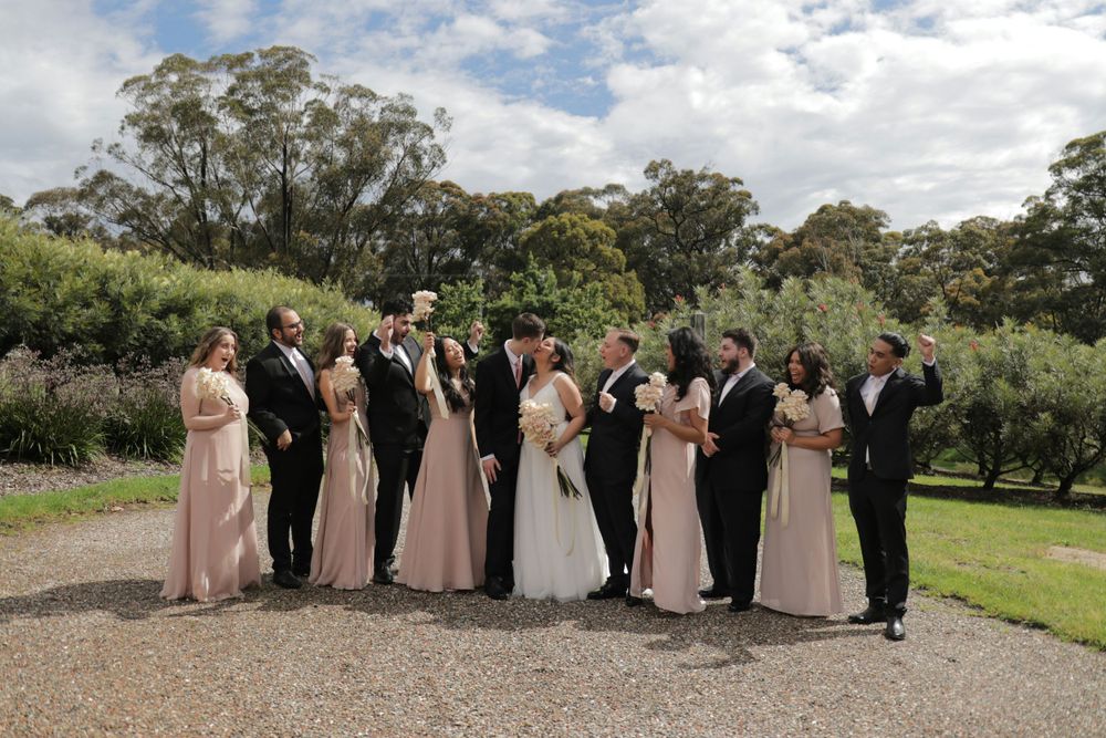 A wedding party in formal attire poses outdoors on a gravel path surrounded by lush green trees and gardens.