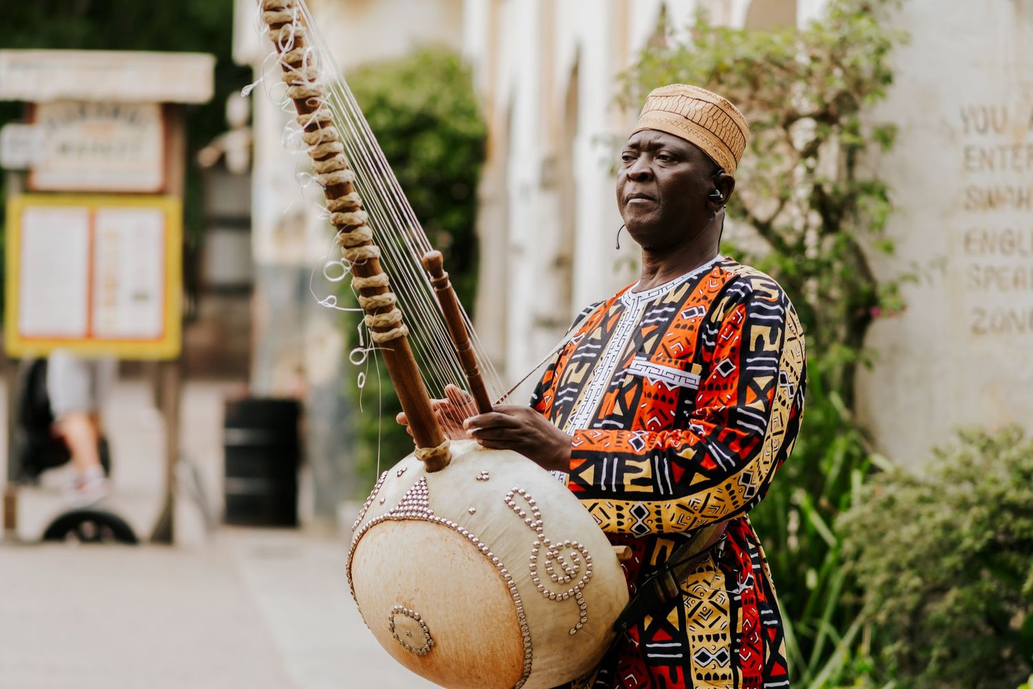 A musician in vibrant African attire plays a traditional kora instrument on a sunny street.