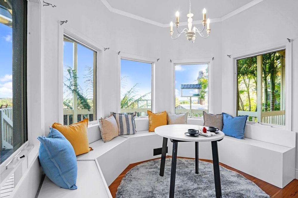 Sunlit curved window seating area with colorful cushions and chandelier in white room.